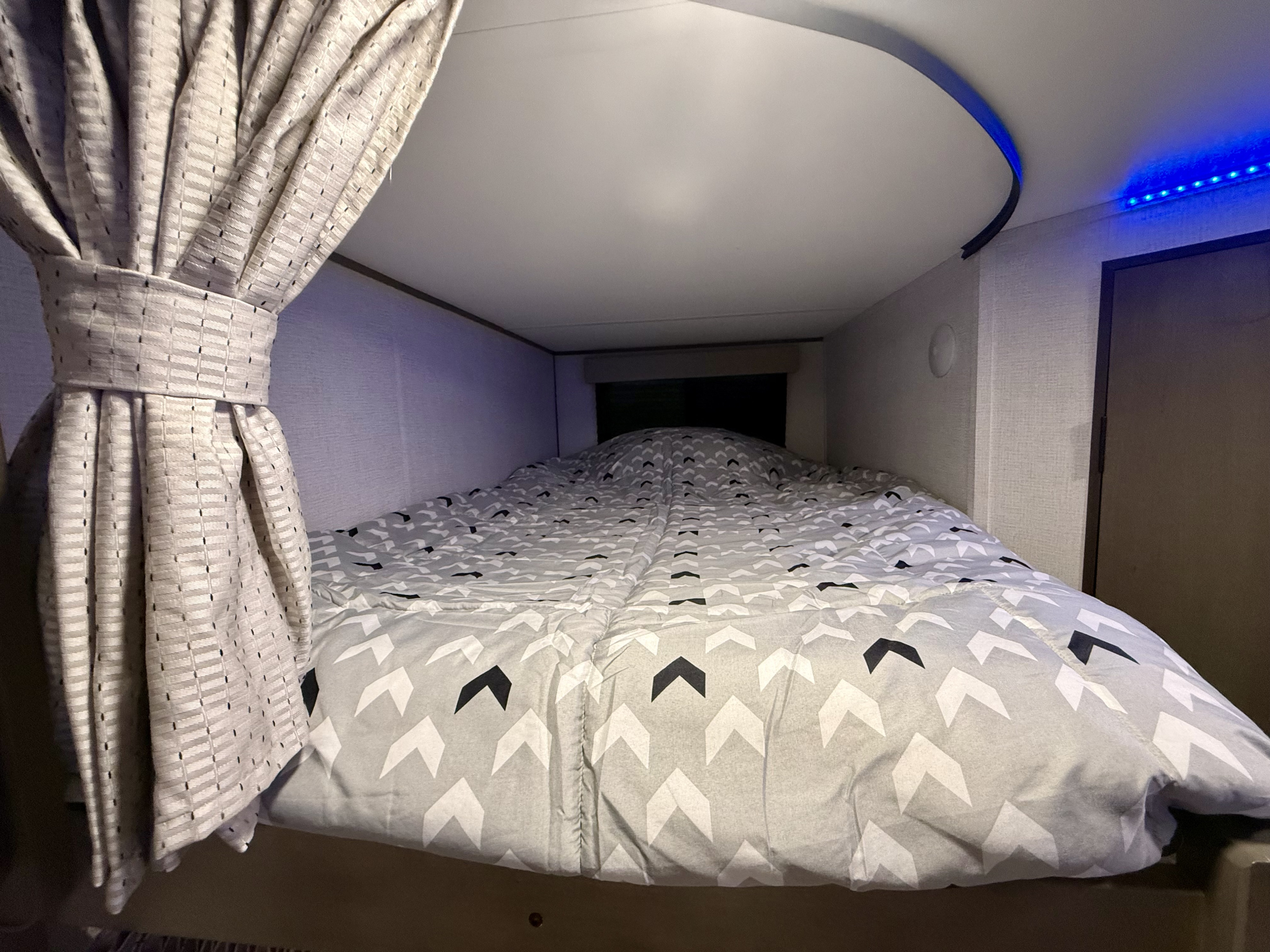 Cozy RV rental bunk bed with grey chevron duvet, tied-back curtain, and blue accent lighting.. Grand Design Transcend Xplor 2025