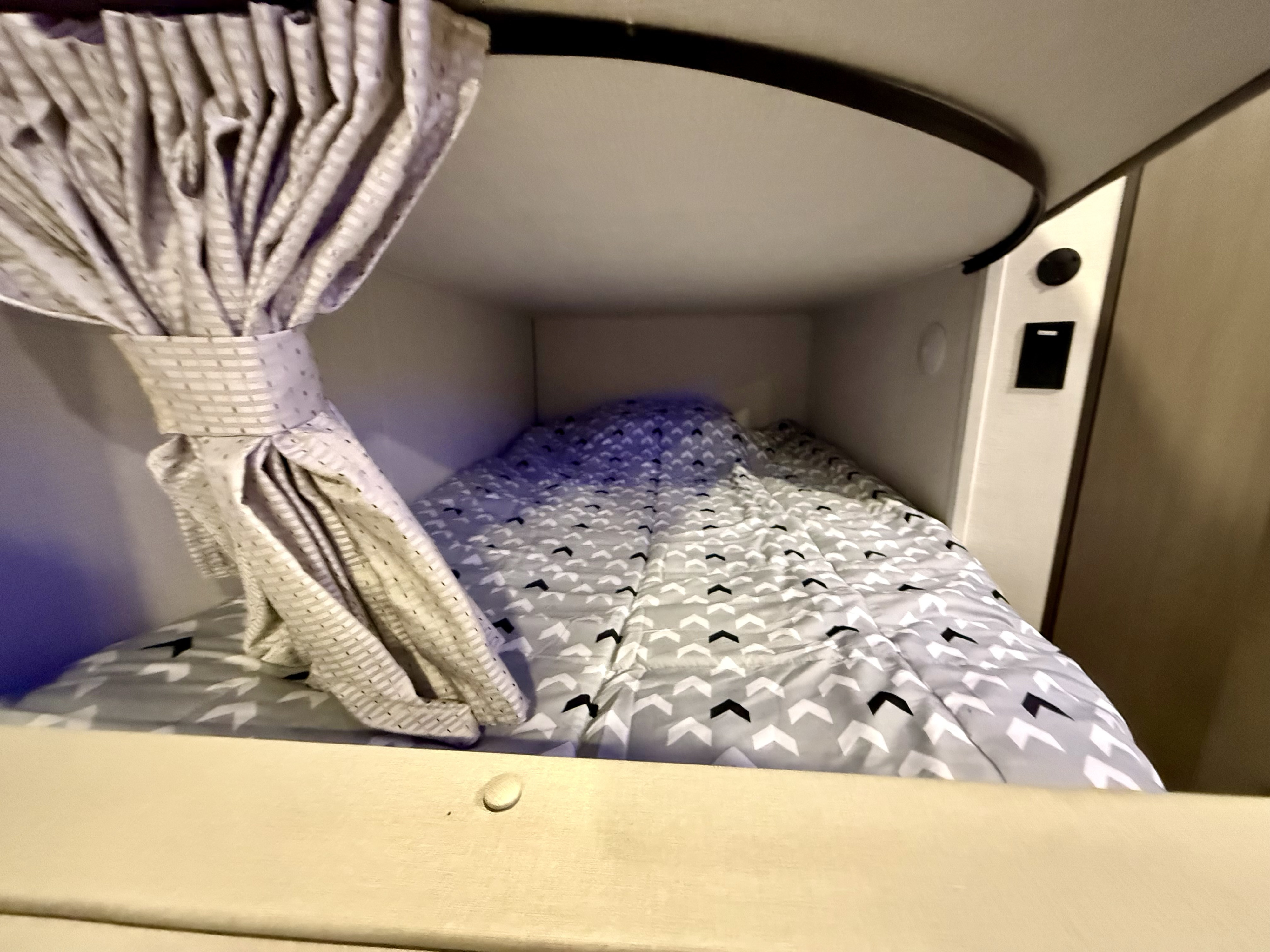Cozy rv rental bunk bed with a grey chevron duvet and tied-back privacy curtain, featuring wall-mounted controls.. Grand Design Transcend Xplor 2025