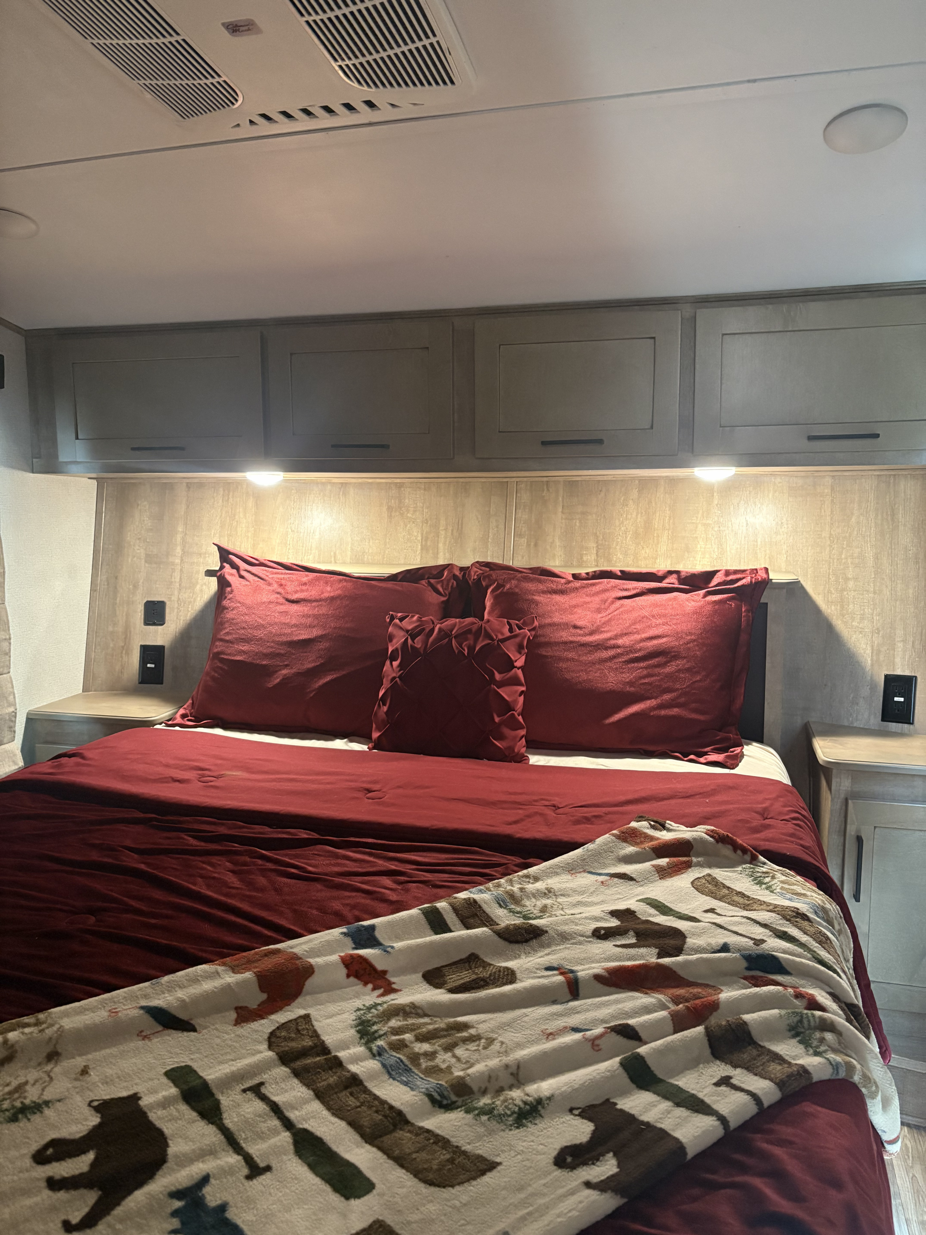 Relax in this cozy RV rental bedroom. Features a comfortable bed with deep red linens, a nature-themed blanket, and overhead storage.. Keystone RV Arcadia 2022