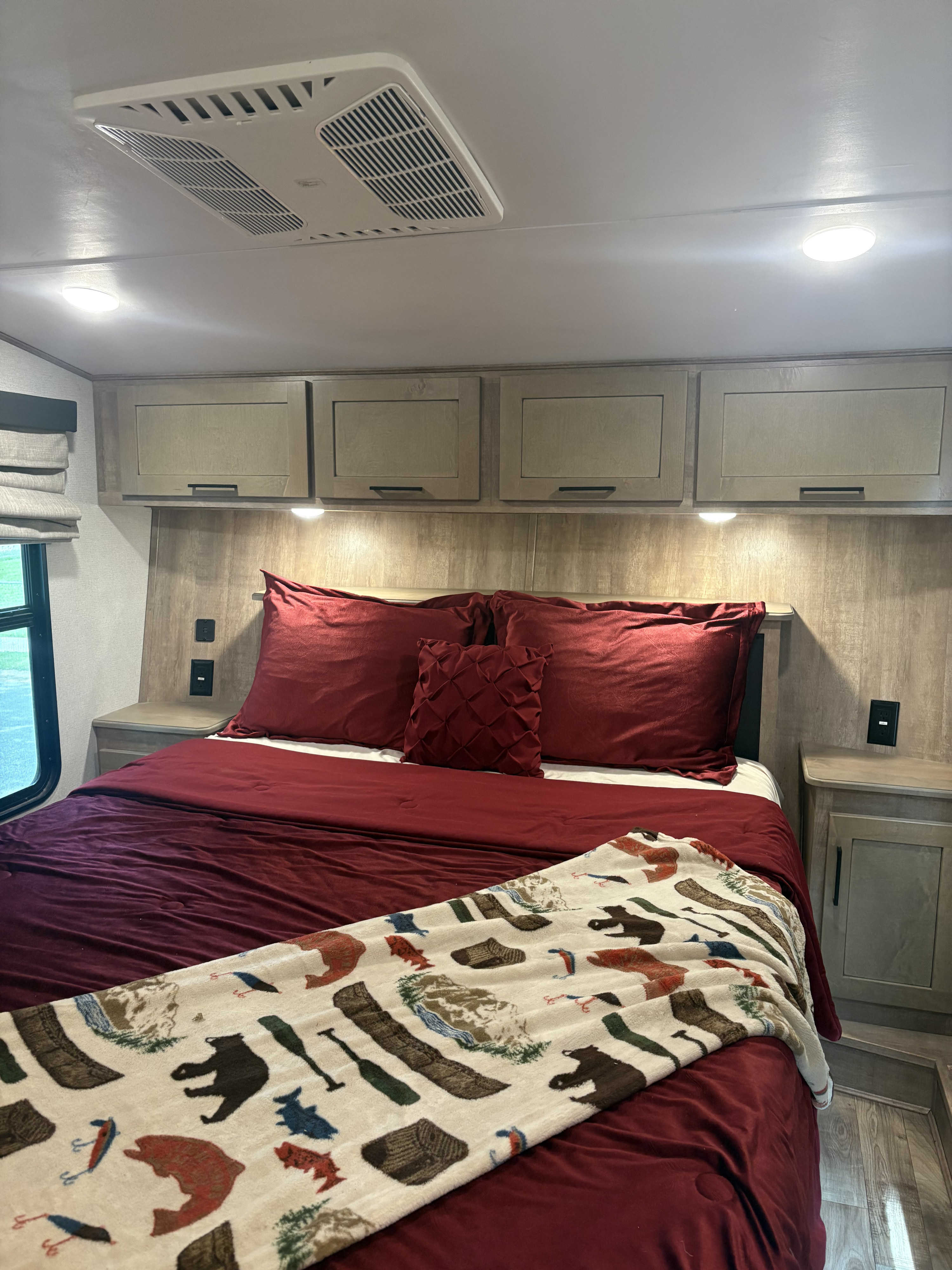 Cozy RV rental bedroom featuring a burgundy bed, nature-themed blanket, overhead cabinets, lights, and AC.. Keystone RV Arcadia 2022