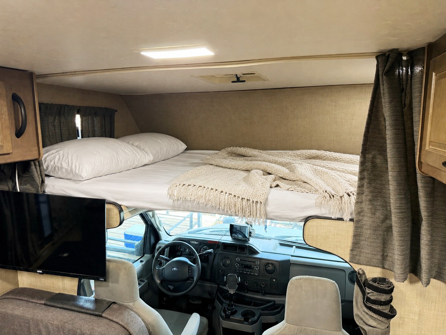 Inside this RV rental, a cozy overhead bed with pillows and blankets is positioned above the driver's cab, alongside a mounted TV.. Thor Majestic 2018