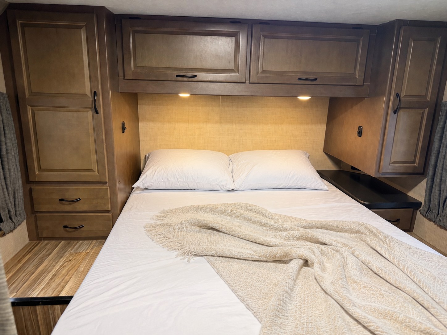 Cozy RV rental bedroom featuring a comfortable bed with white linens, wooden storage cabinets, and warm overhead lighting.. Thor Majestic 2018