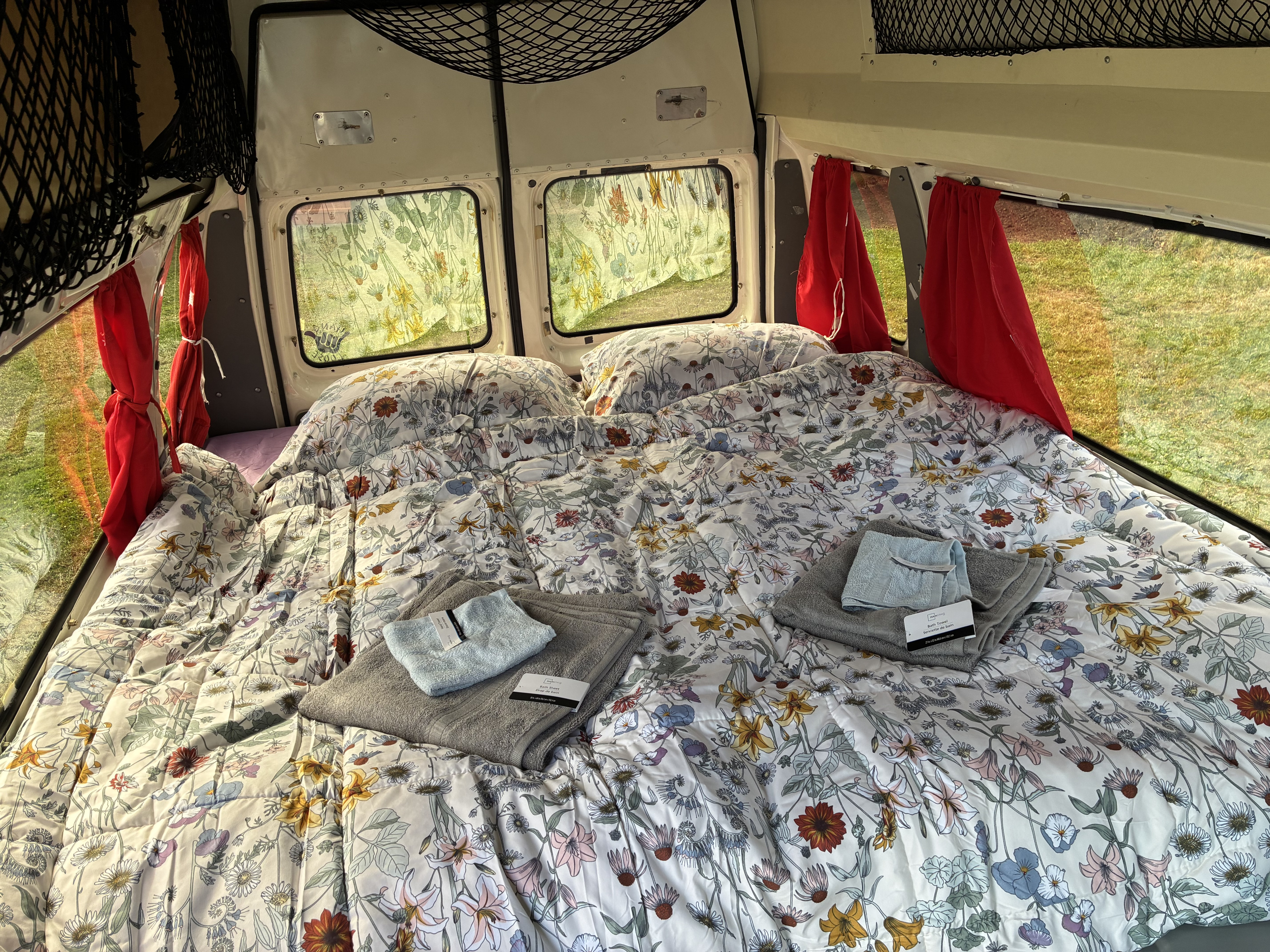 Cozy RV rental bedroom featuring floral bedding, pillows, fresh towels, and sunny windows with red curtains.. Ford Econoline E250supercab 1996