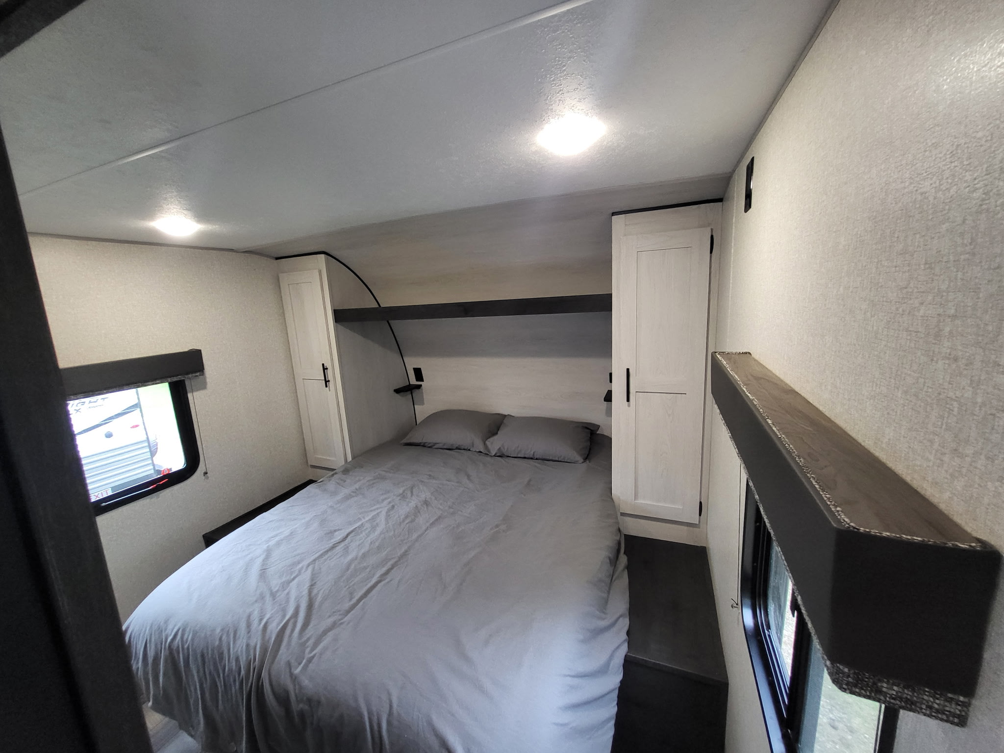 This RV rental offers a cozy bedroom with a neatly made gray bed, flanking cabinets, an overhead shelf, and windows.. KZ Sportsmen SE 332bkhse 2022