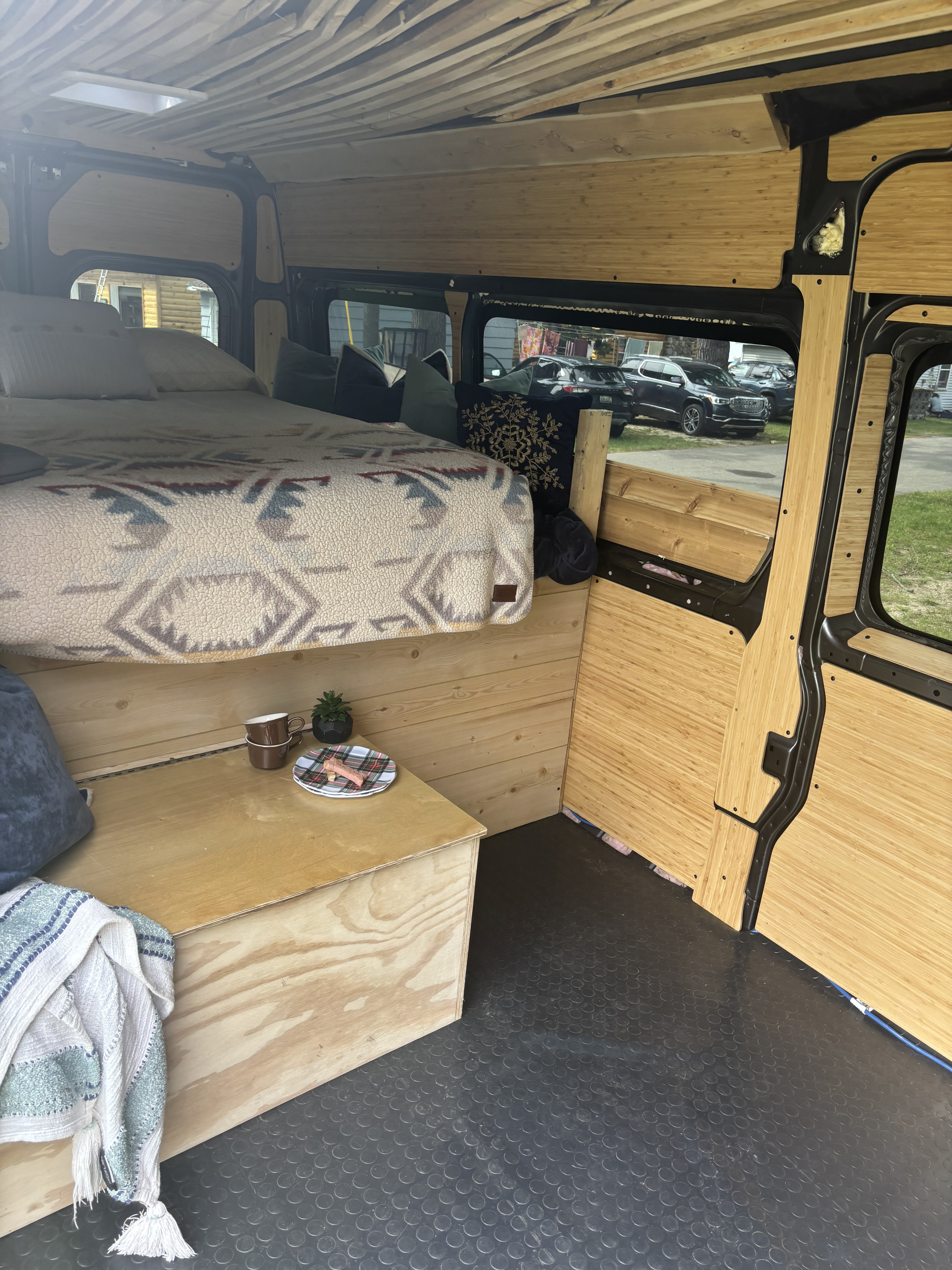 Interior of an RV rental camper van, featuring a raised bed with a blanket, wooden walls & ceiling, and rubber flooring.. Dodge Promaster 2023