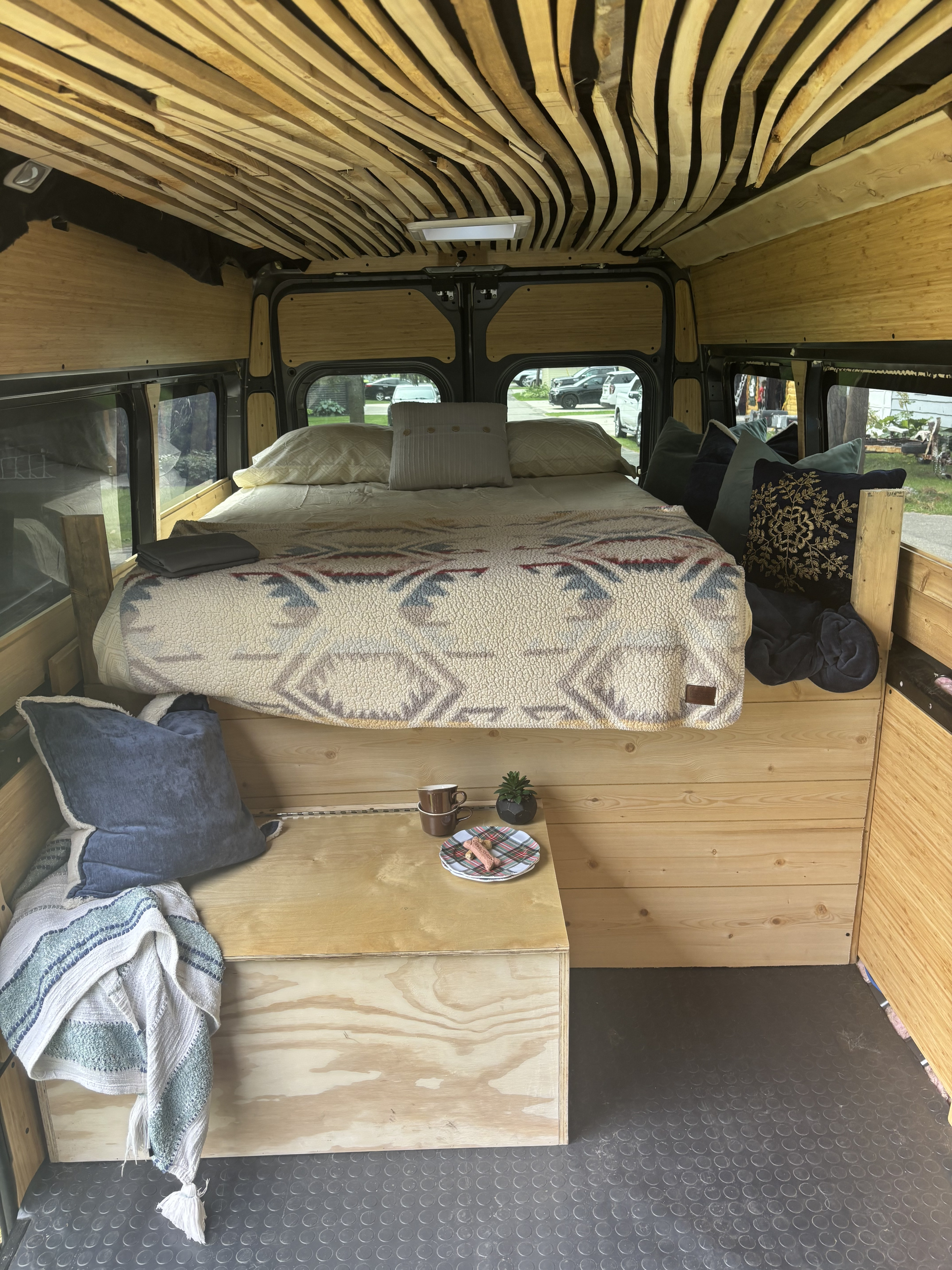 This cozy rv rental camper van features a unique wooden slat ceiling, a comfortable bed, and a small table with snacks.. Dodge Promaster 2023