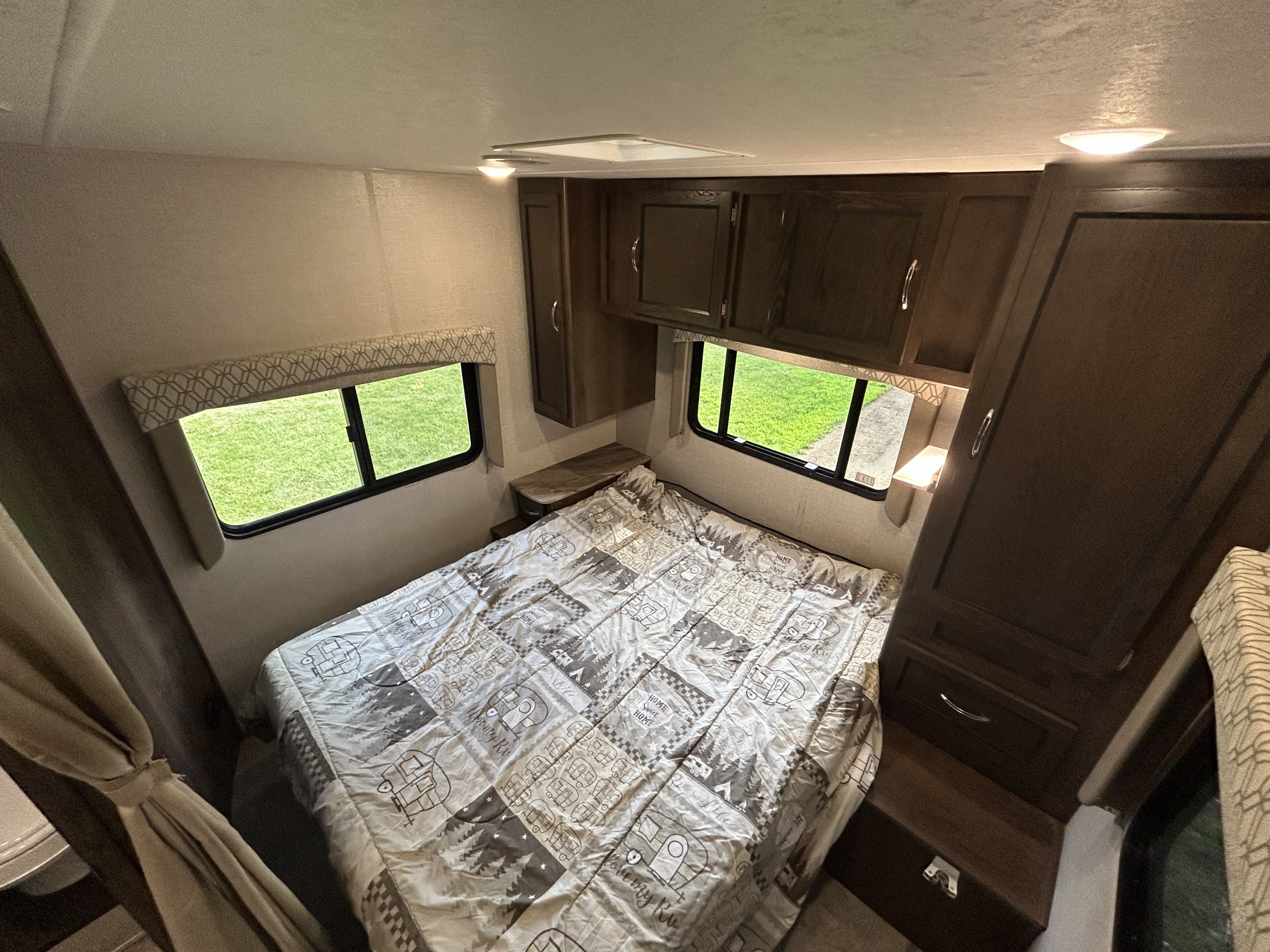 Explore this RV rental's cozy bedroom featuring a patterned comforter, overhead cabinets, and bright windows with green views.. Coachman RV 27QB Chevy 4500 2020