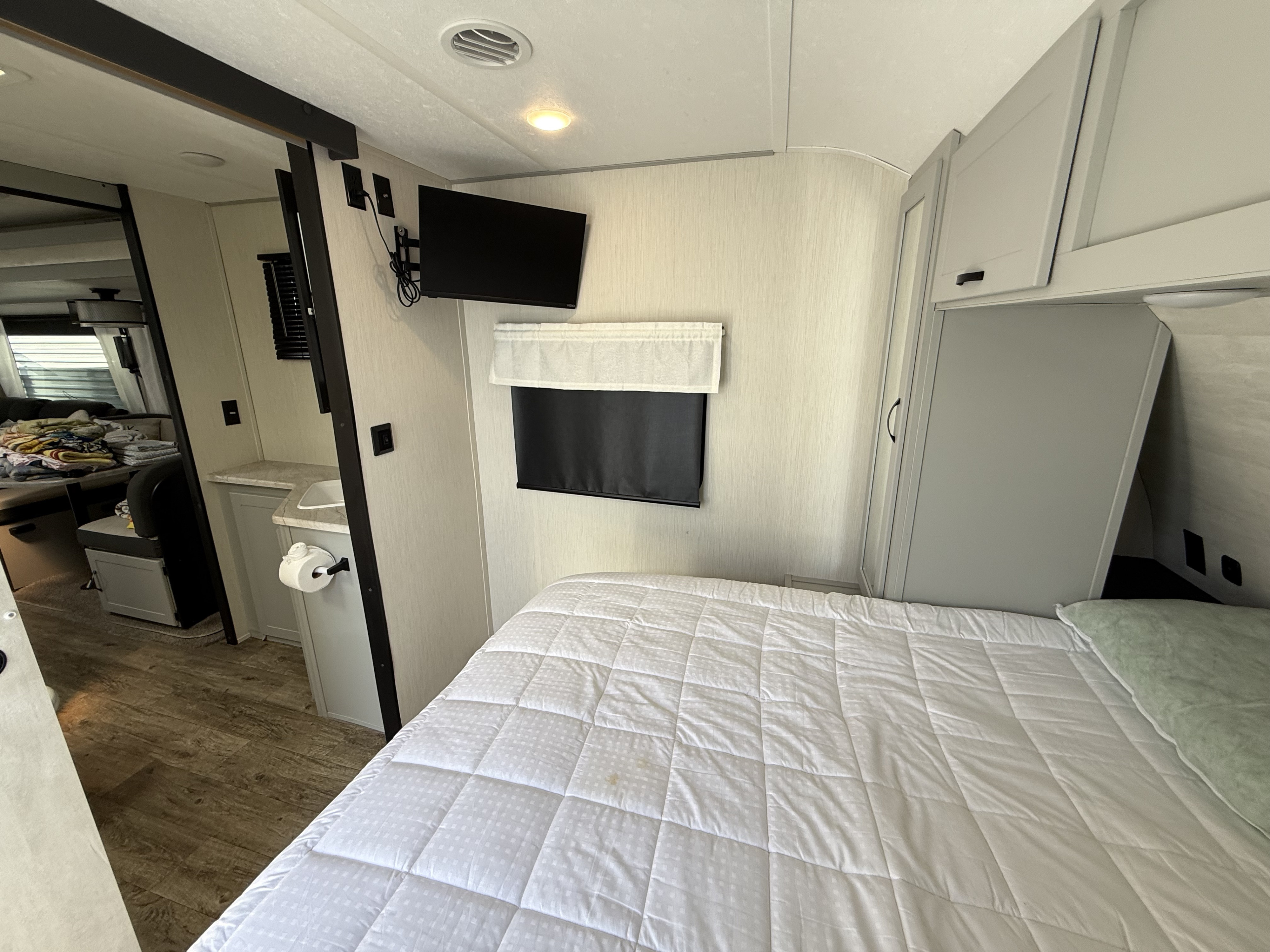 This RV rental bedroom features a comfy white bed, wall-mounted TV, storage cabinets, and access to a compact bathroom.. Forest River Vibe 2023