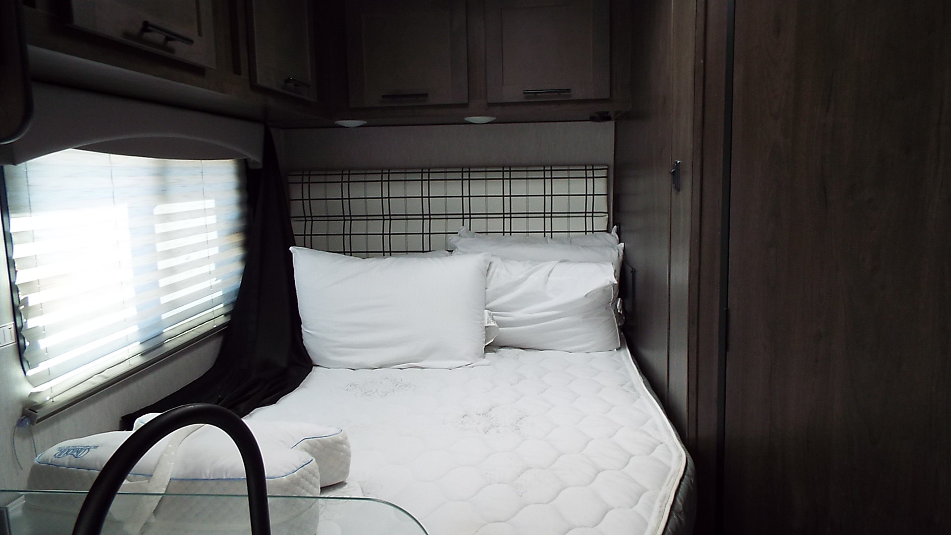 Inside an RV rental, this bedroom features a bed with white pillows, a plaid headboard, overhead cabinets, and a bright window.. Coachmen Ford Transit 2023