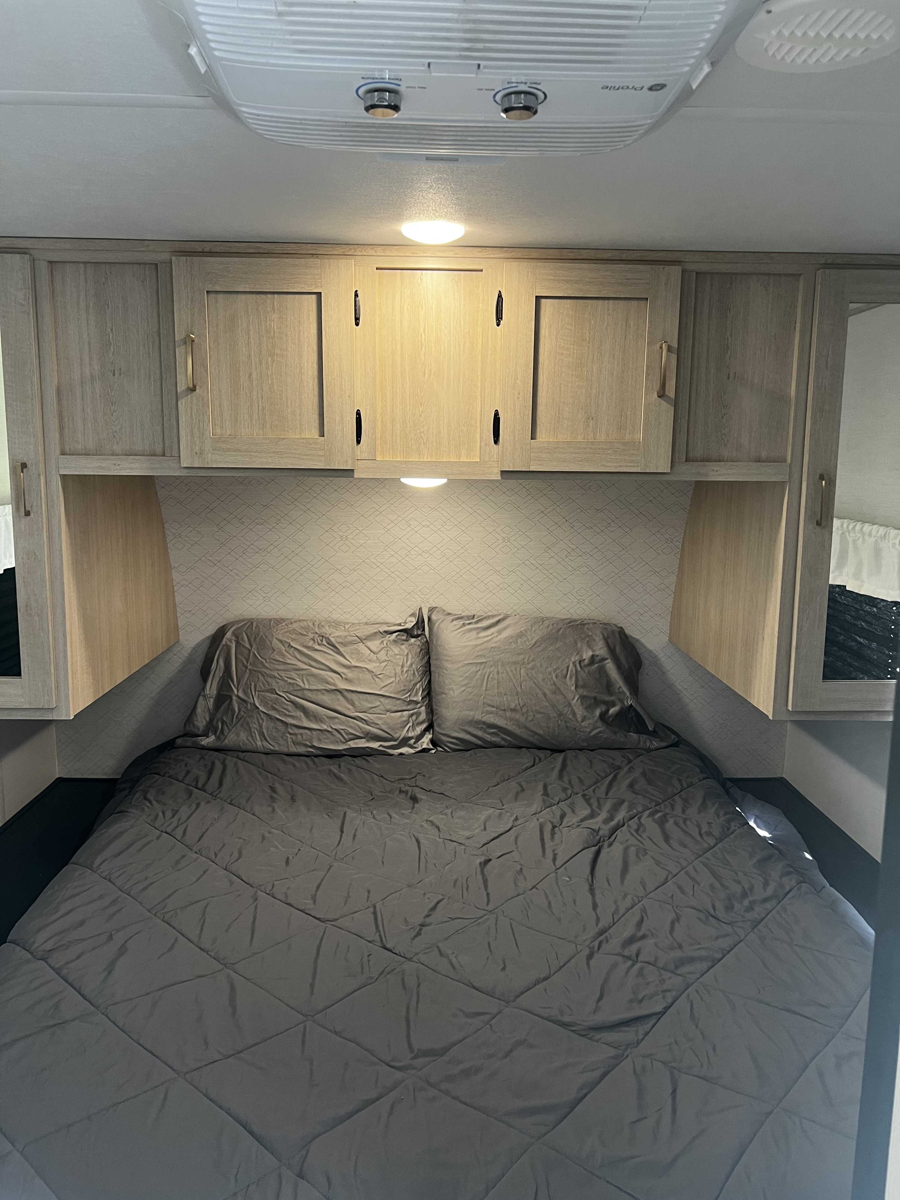 This RV rental bedroom features a comfy gray bed with pillows, overhead wooden cabinets, and a ceiling air conditioner for comfort.. Avenger Avenger Trailer 2025