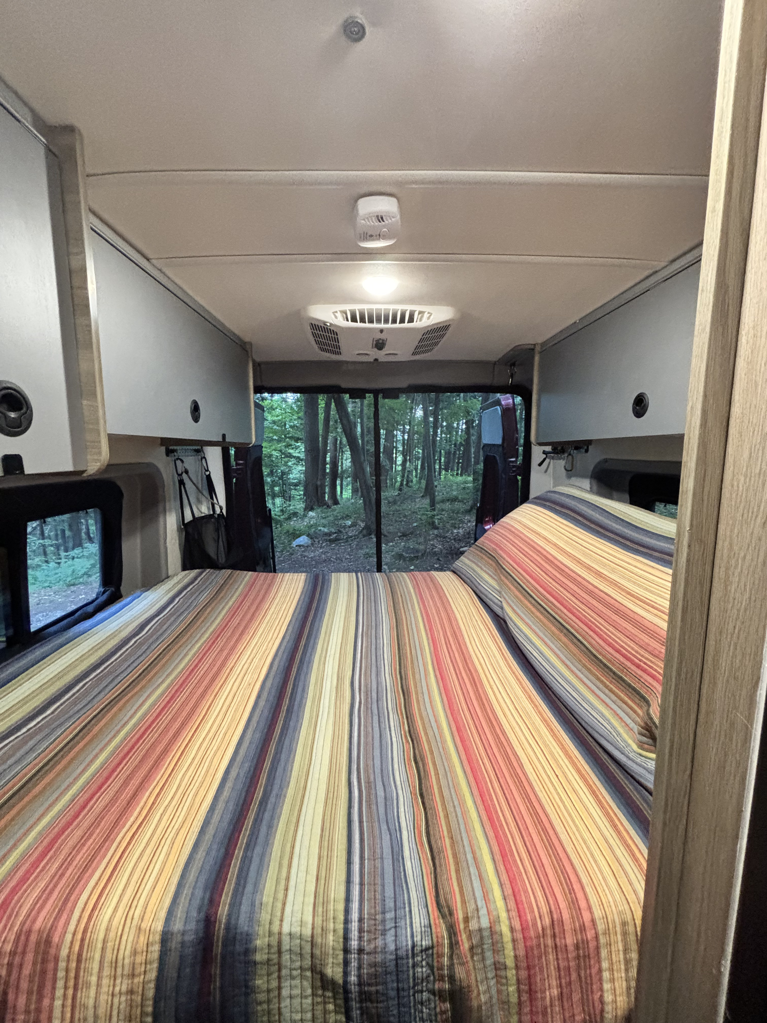 Cozy rv rental interior features a colorful striped bed, overhead storage, and a forest view from the rear.. Dodge Winnebago 2021