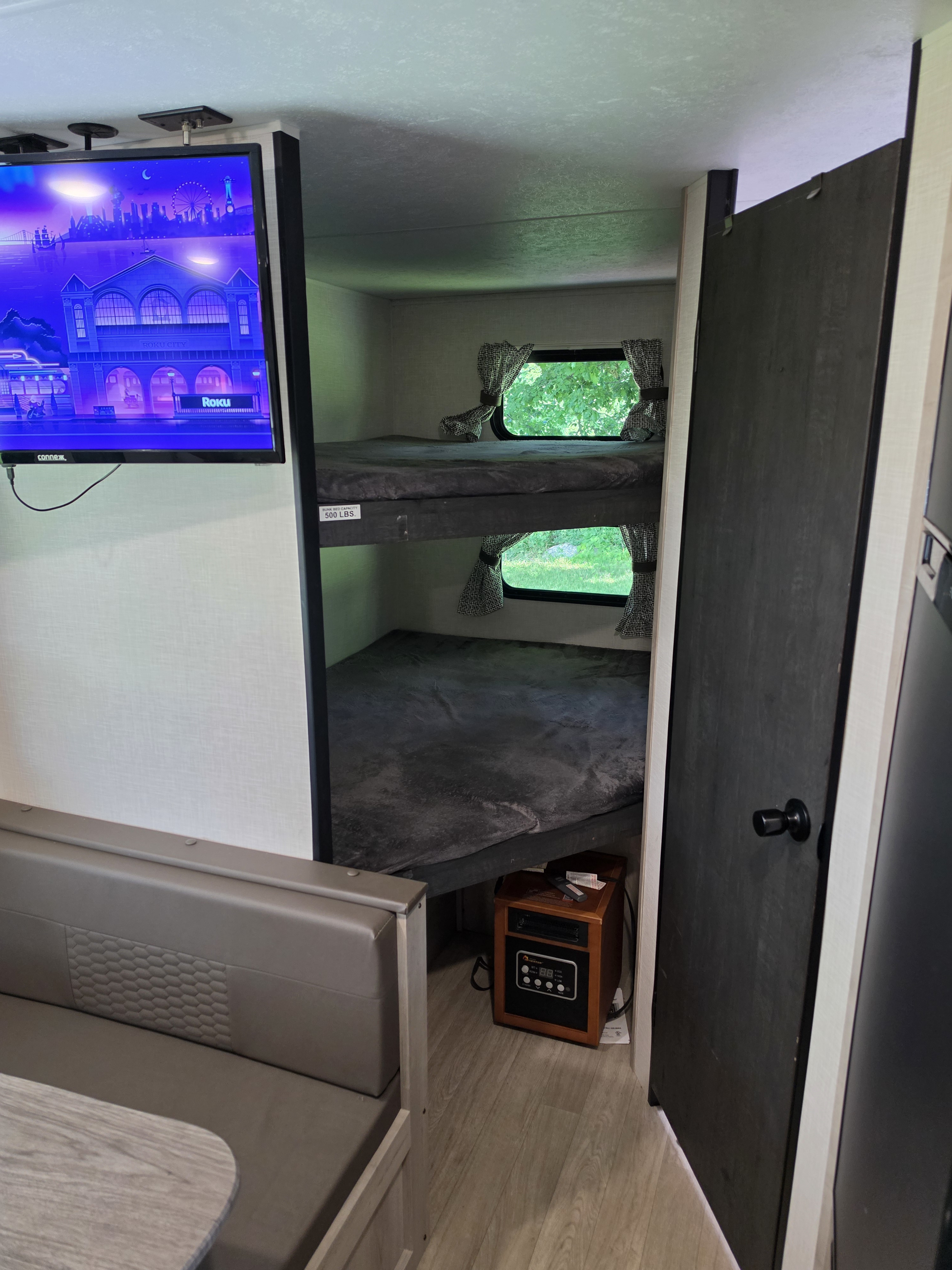 Inside an RV rental, showing bunk beds with windows, a TV, and a dining area. An electric heater sits below the bunks.. KZ Sportsmen 2024