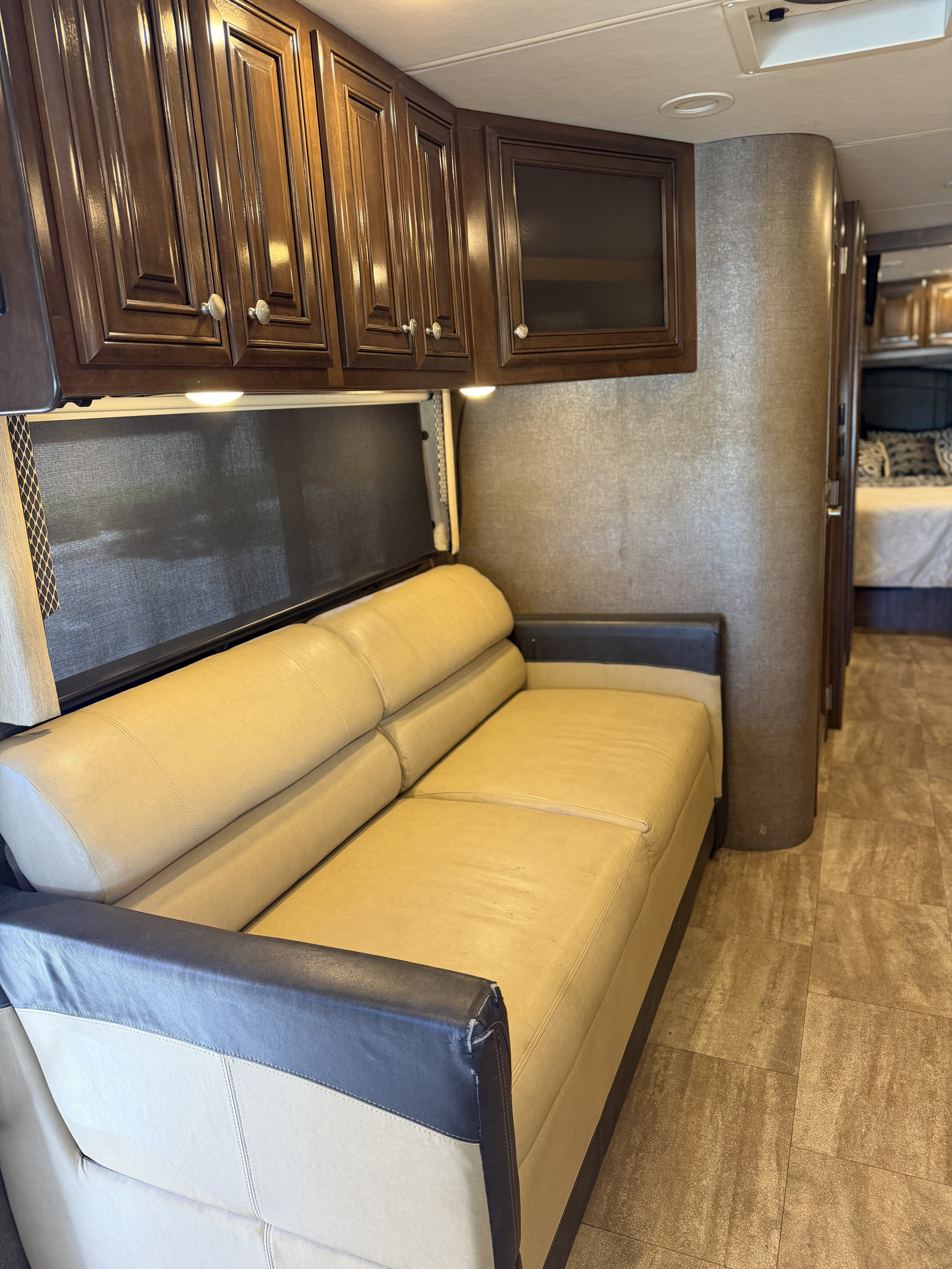 RV rental interior features a cozy beige sofa, rich wood cabinets, and patterned tile floor, with a bedroom visible in the background.. Thor Palazzo 2017