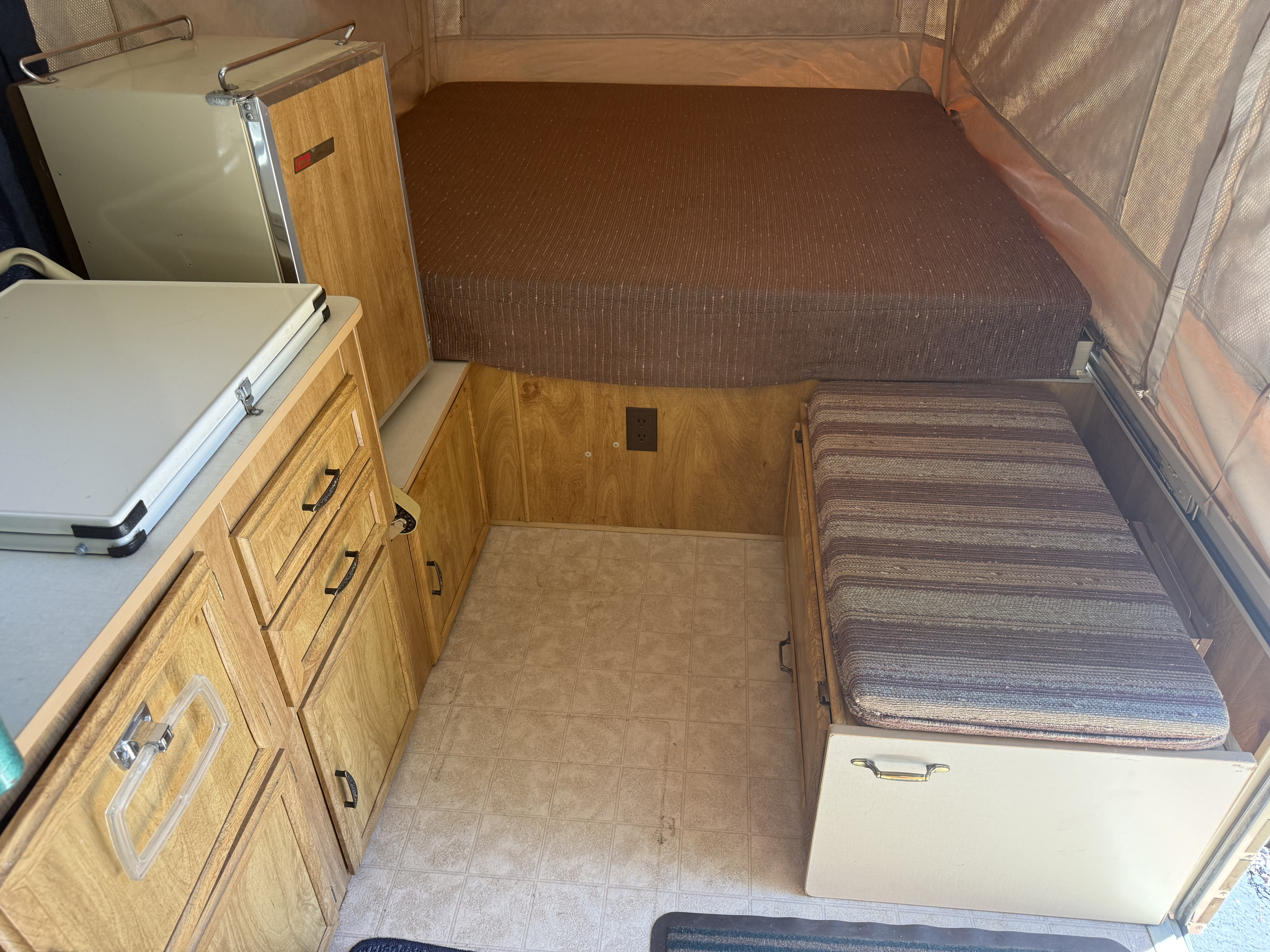 RV rental pop-up camper interior: a large bed, kitchenette with wood cabinets, and bench seating with storage.. Coleman Grand Tour Shenandoah 1987