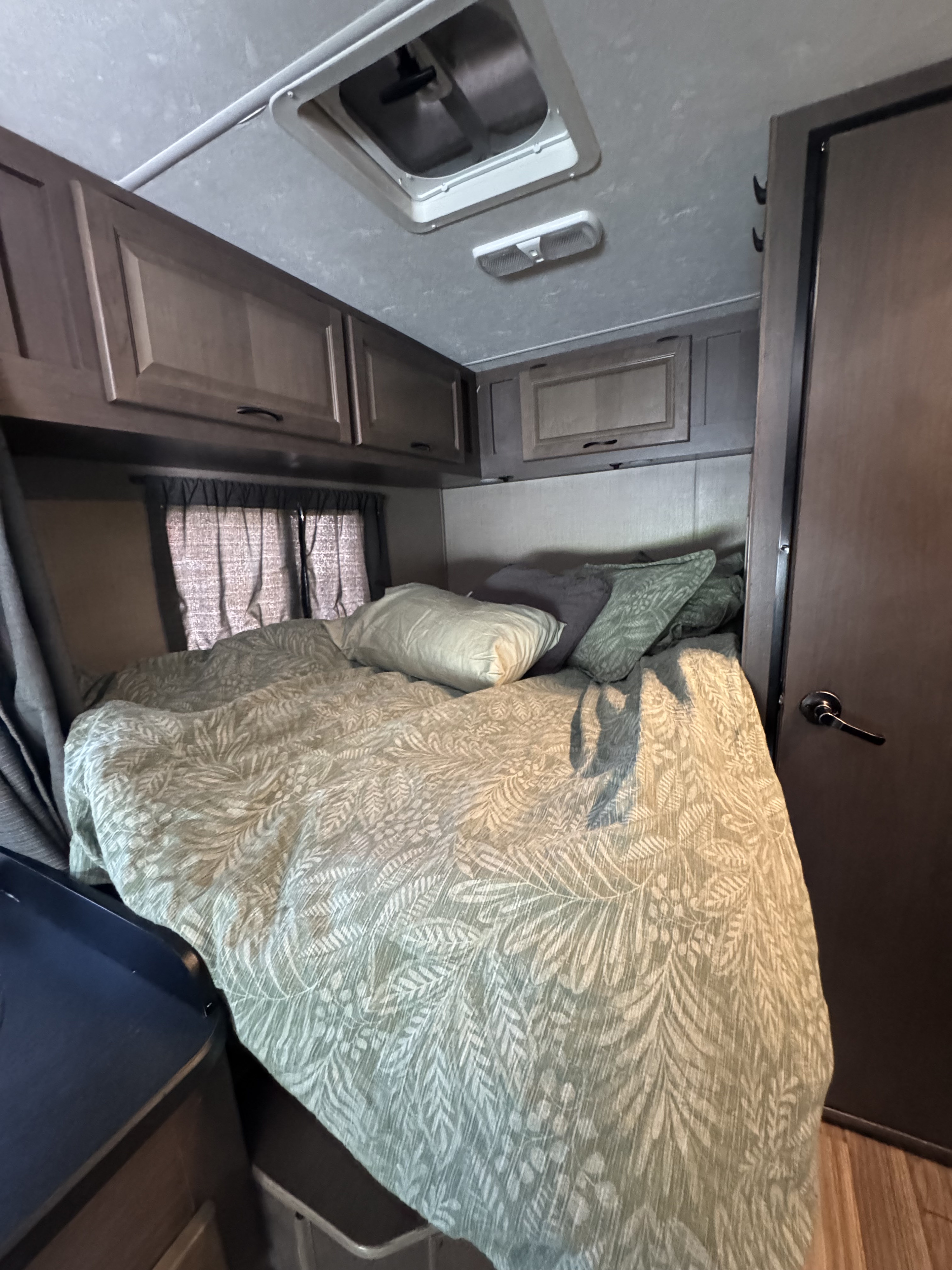 Compact RV rental bedroom featuring a comfy bed with green botanical bedding, dark wood cabinets, and overhead vent.. Thor Majestic 2018