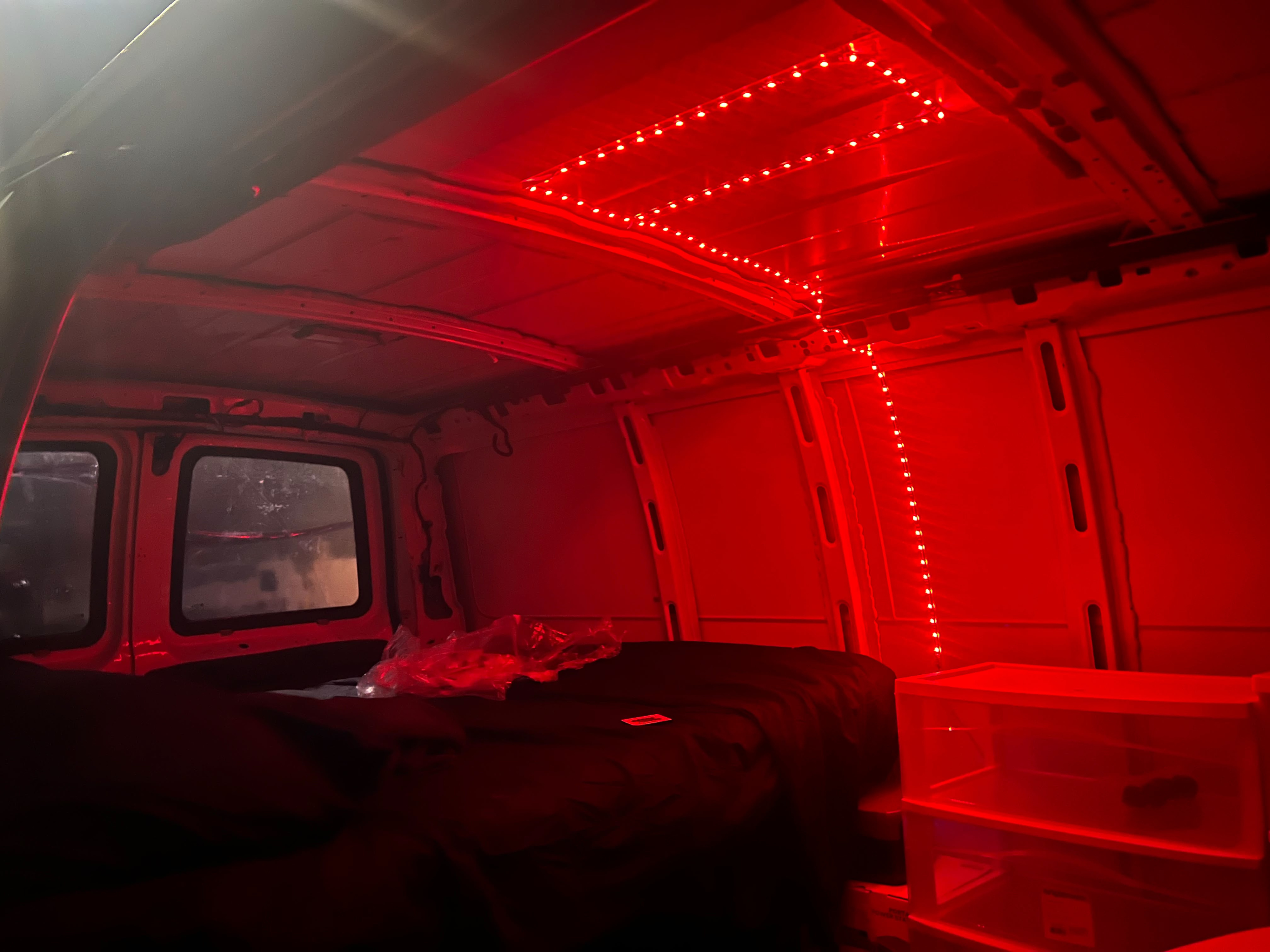 RV rental interior: van bathed in red LED string lights on ceiling & walls, illuminating a dark bed and white plastic drawers.. Chevrolet Express 2006