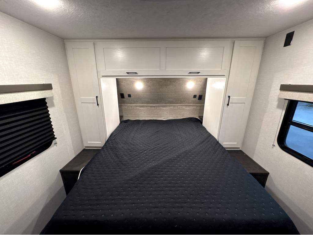 RV rental bedroom showcasing a bed with dark quilt, white overhead cabinets, side wardrobes, and windows with blinds.. Sienna SA281VQB 2025