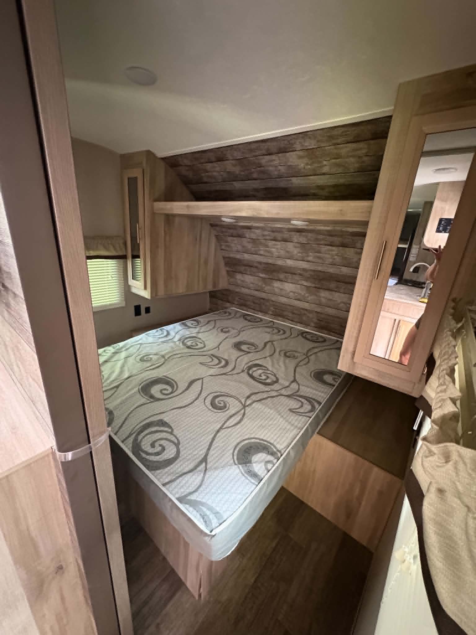 RV rental bedroom with patterned mattress, wood-paneled walls, built-in storage, and a mirrored cabinet reflecting the kitchen.. Coachmen Catalina Summit 261bhs 2021
