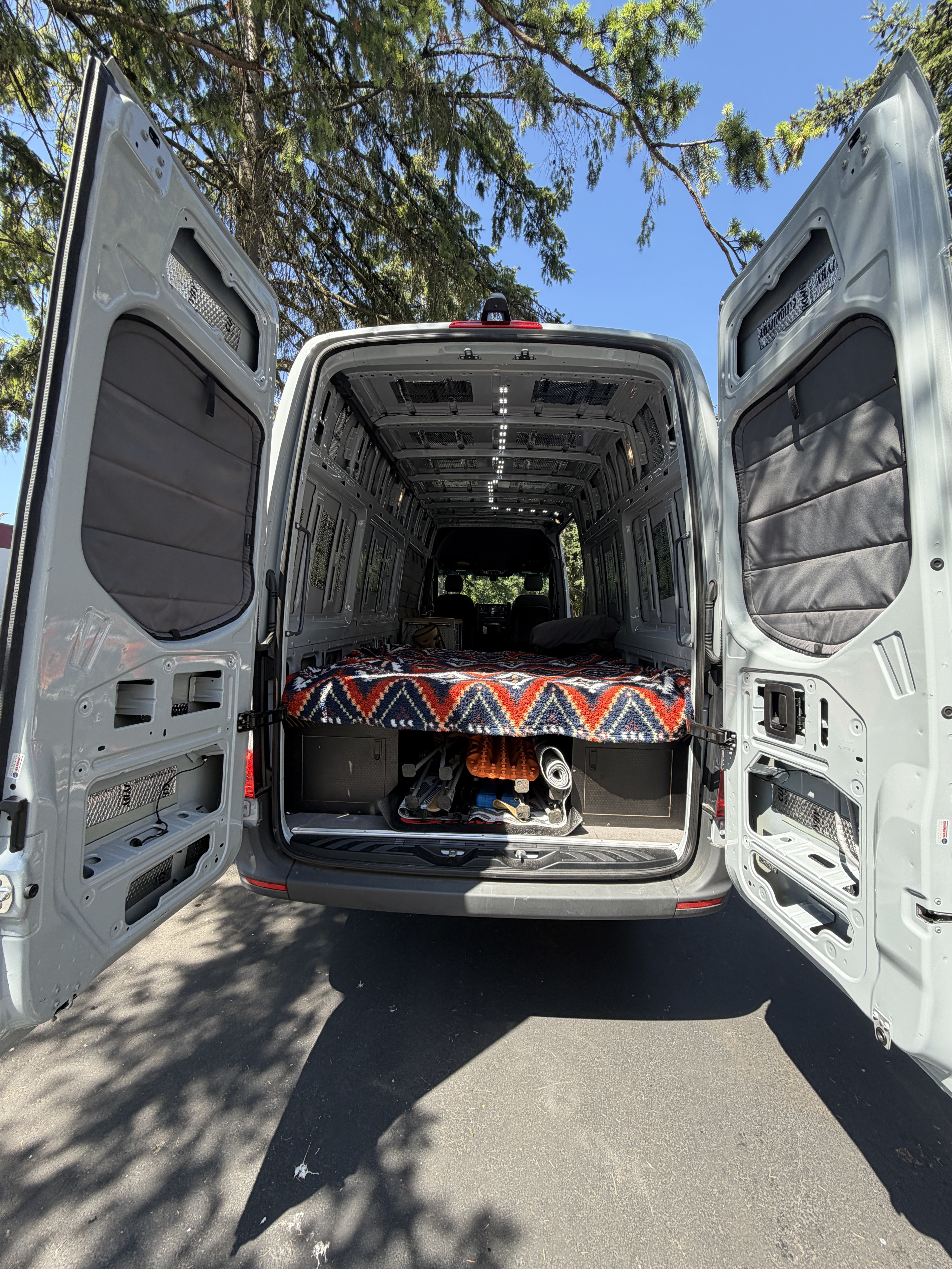 Open rear of an RV rental camper van shows a partially converted interior with bed, under-bed storage, and sound-insulated walls.. Mercedes Sprinter 2500 2025