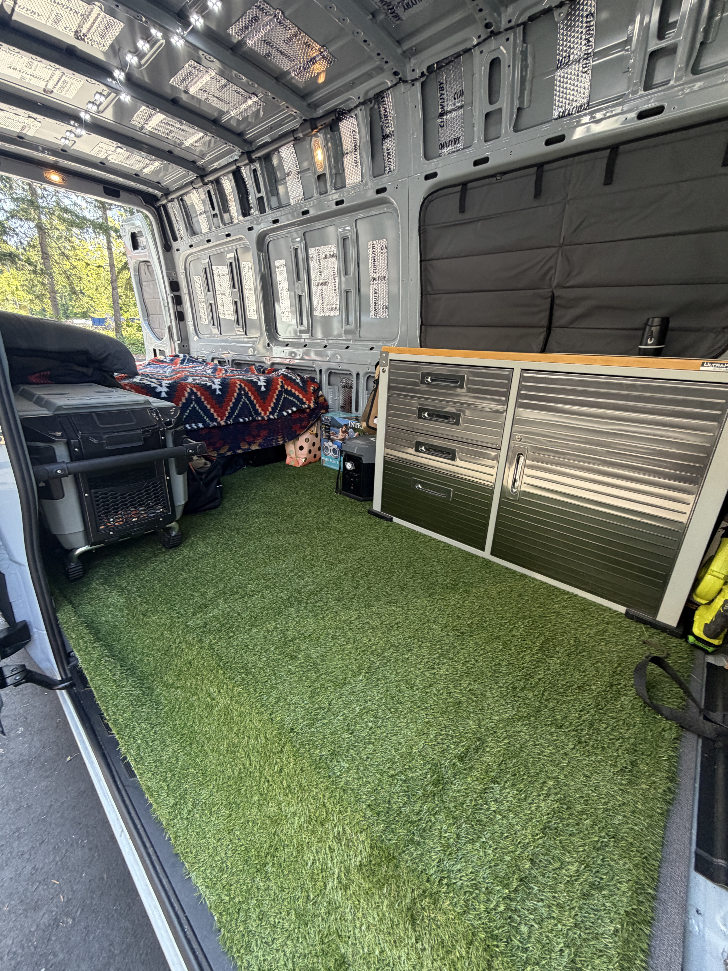 Inside a DIY rv rental campervan: artificial turf floor, cozy bed, fridge, and metal storage. Walls have sound deadening.. Mercedes Sprinter 2500 2025