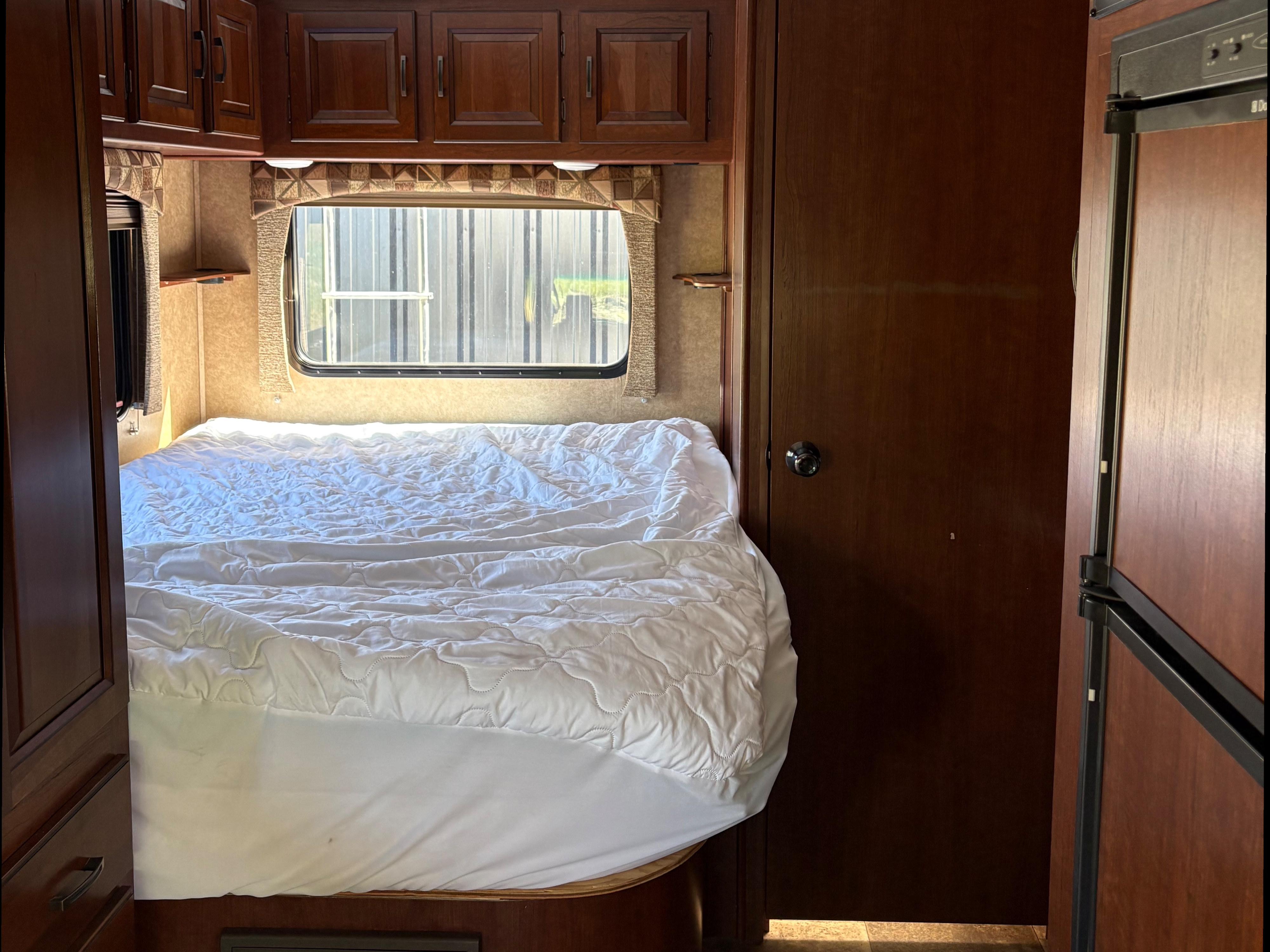 RV rental bedroom: a cozy bed with white bedding, overhead wooden cabinets, a window, and part of a built-in fridge.. Forest River Sunseeker 2015