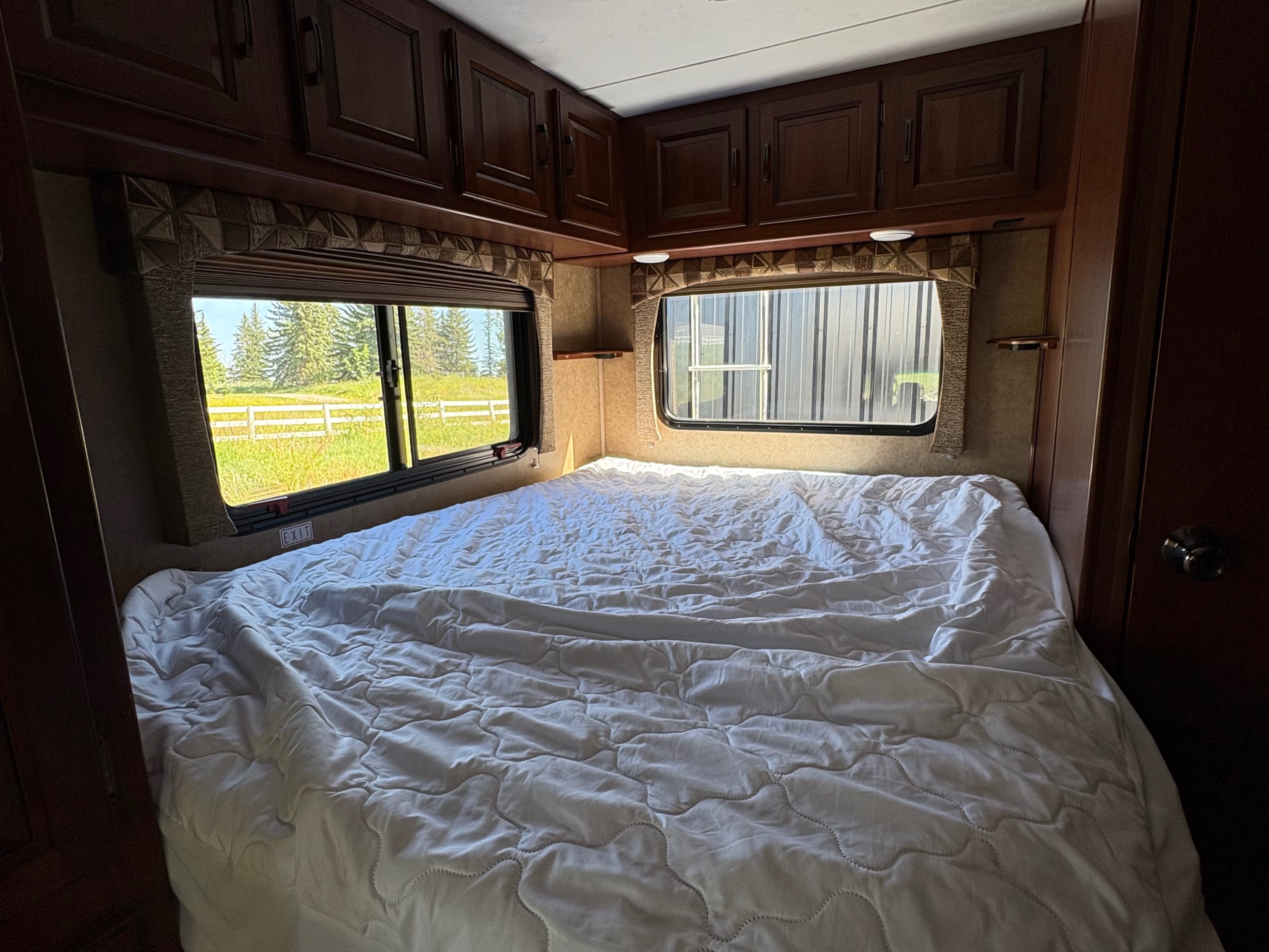 Cozy RV rental bedroom featuring a large white bed, windows with outdoor views of trees and a fence, and overhead cabinets.. Forest River Sunseeker 2015