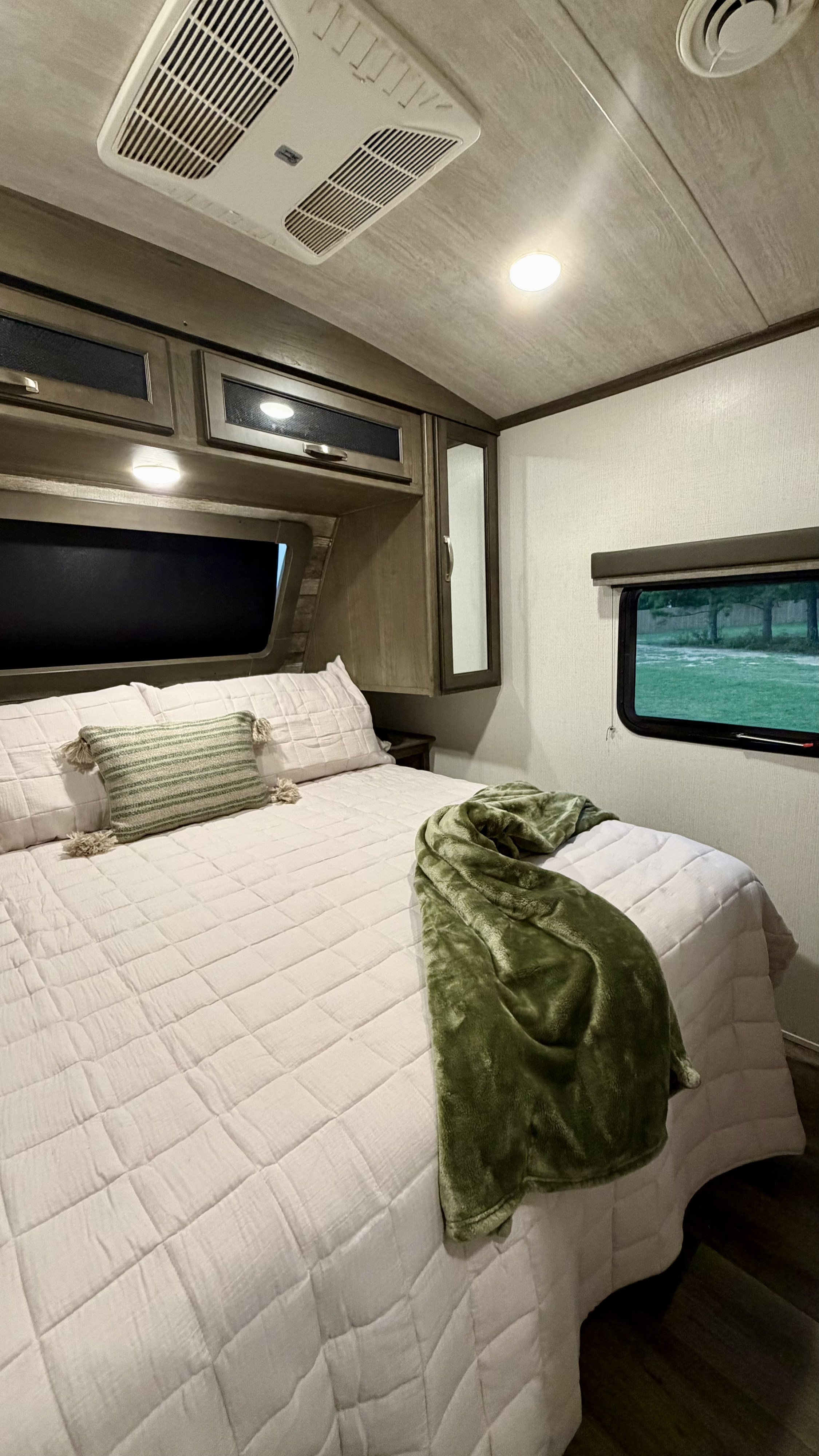 Step into this comfortable RV rental bedroom, featuring a queen bed with light bedding, overhead cabinets, and a window with a green view.. Keystone RV Cougar 2021