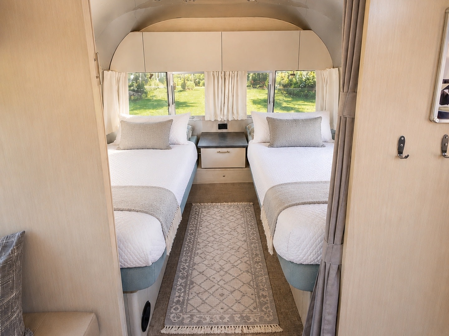 Relax in this RV rental bedroom with two cozy twin beds, white linens, and soft grey accents, offering serene outdoor views.. Airstream Flying Cloud 2025