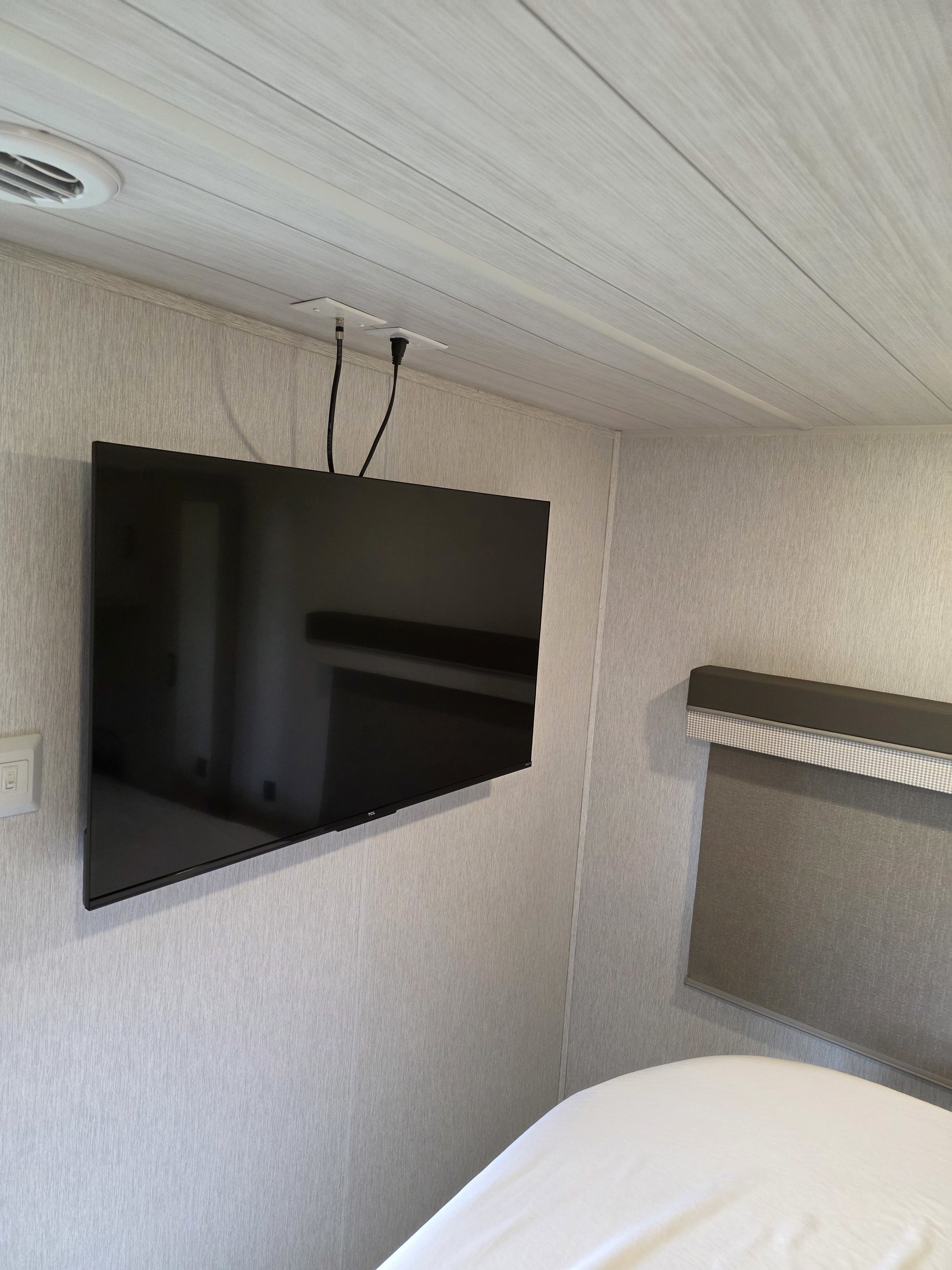 Inside an RV rental, a wall-mounted TV with ceiling-fed cables is shown in a bedroom area with a window blind and white bed sheets.. Forest River Wildwood 2023