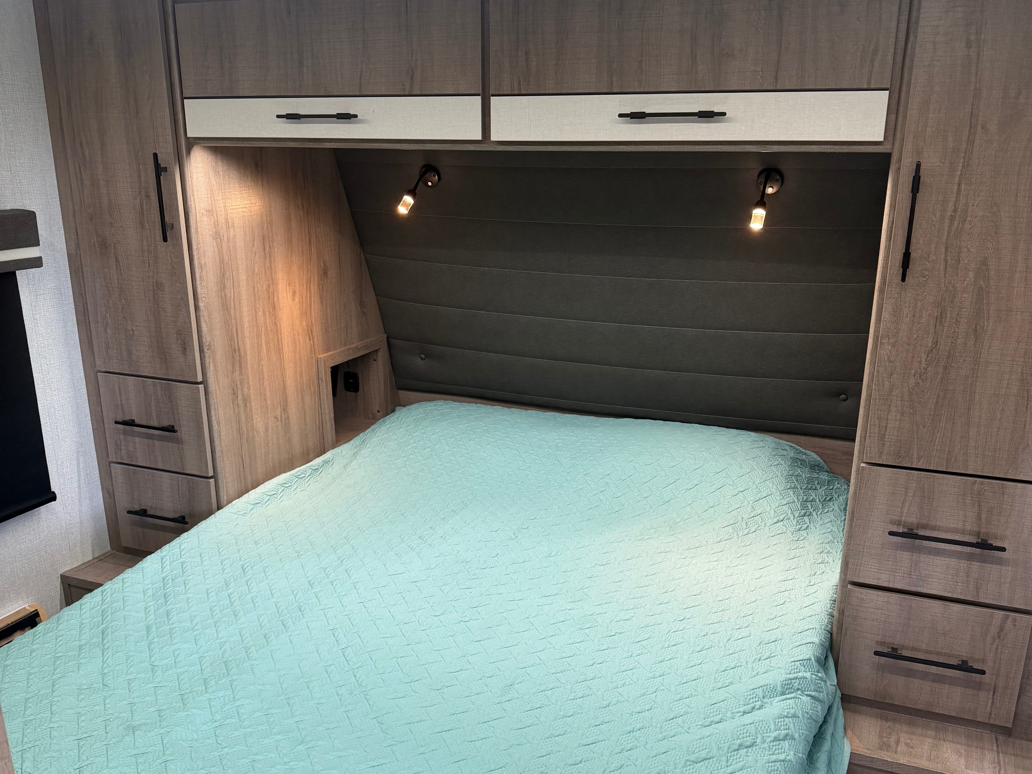 Cozy RV rental bedroom with a teal quilted bed, reading lights, and built-in wood-grain cabinetry, perfect for travel.. Grand Design Imagine XLS 2024