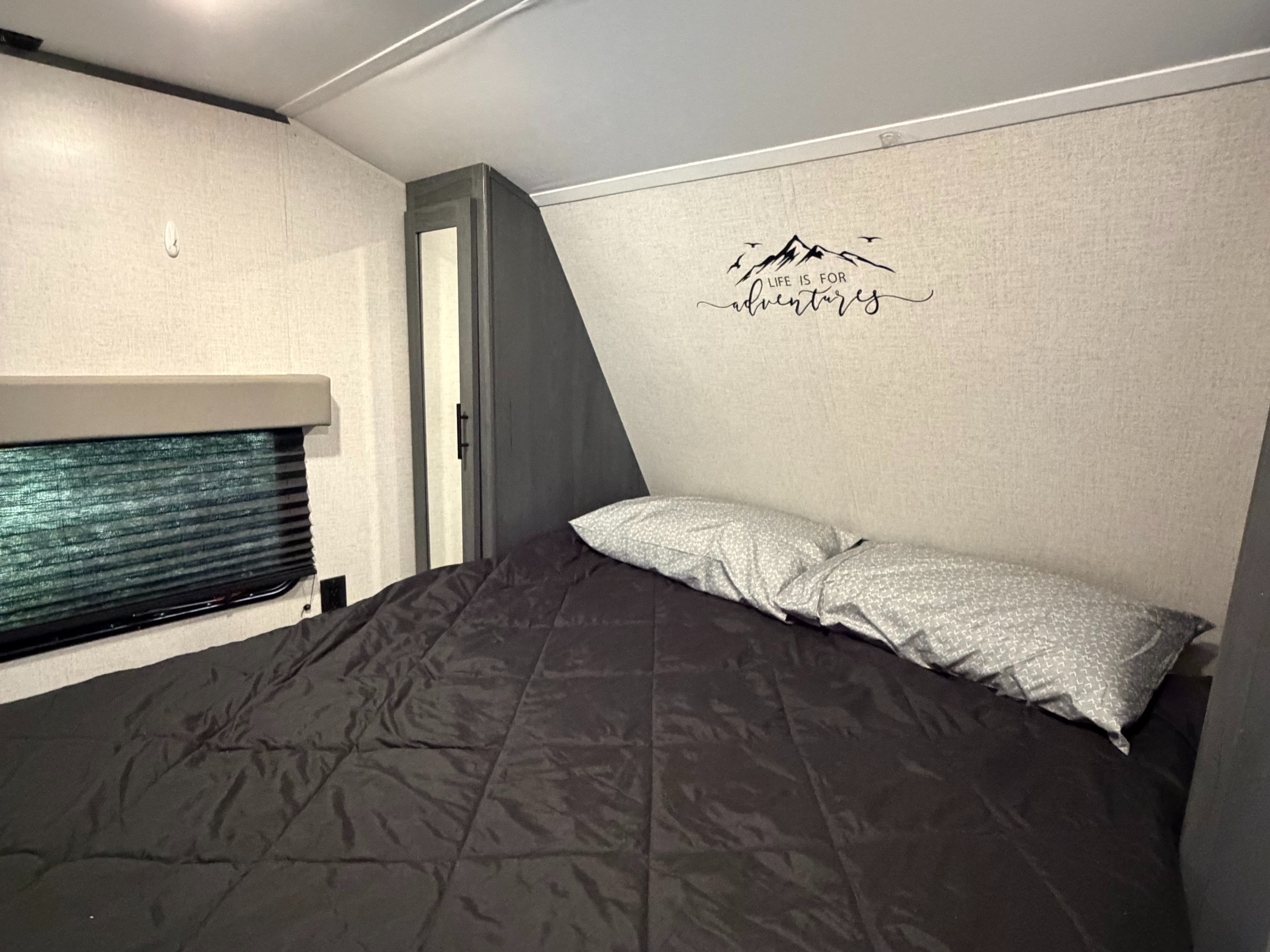 Inside an RV rental, this cozy bedroom features a black comforter, patterned pillows, and a "LIFE IS FOR adventures" wall decal.. Dutchmen Aspen Trail LE 26BH 2022