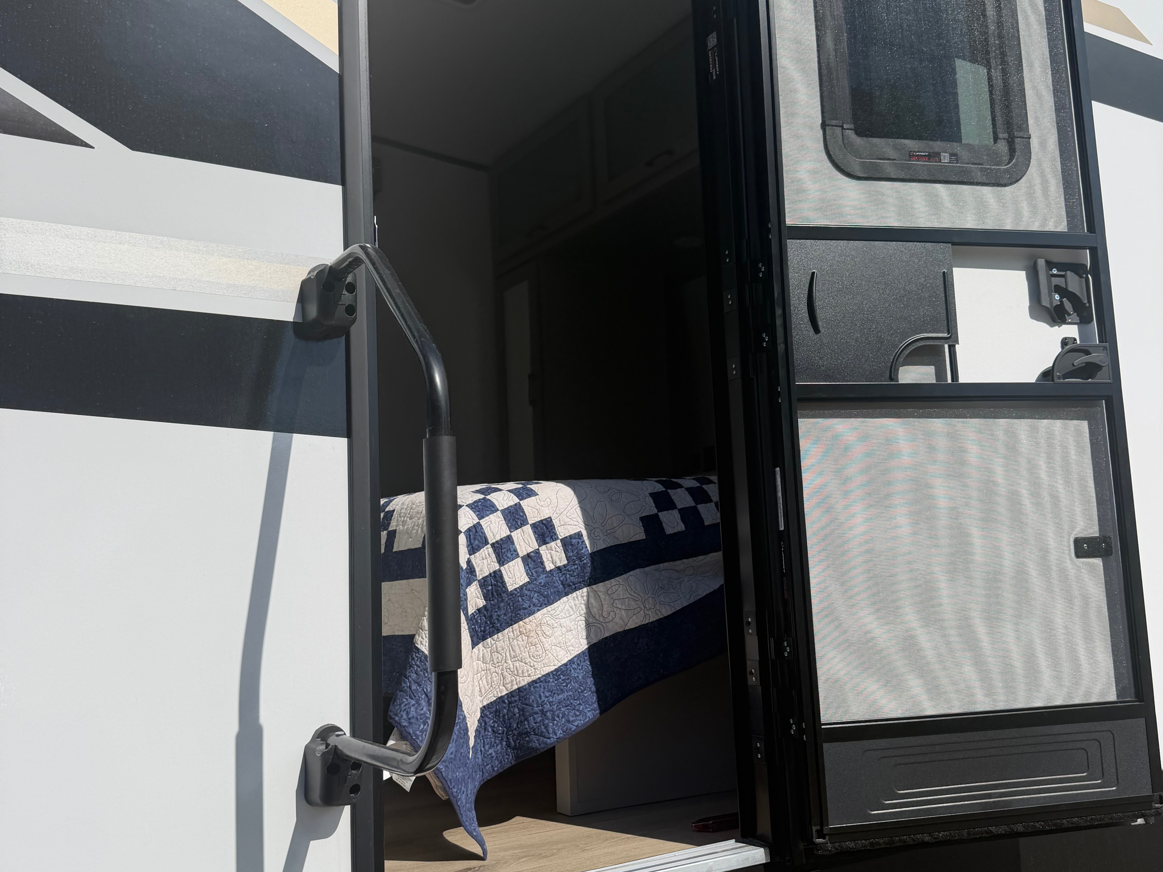 Open door to an RV rental showing a grab handle, screen door, and a bed with a blue and white quilt inside.. Forest River Stealth 2026