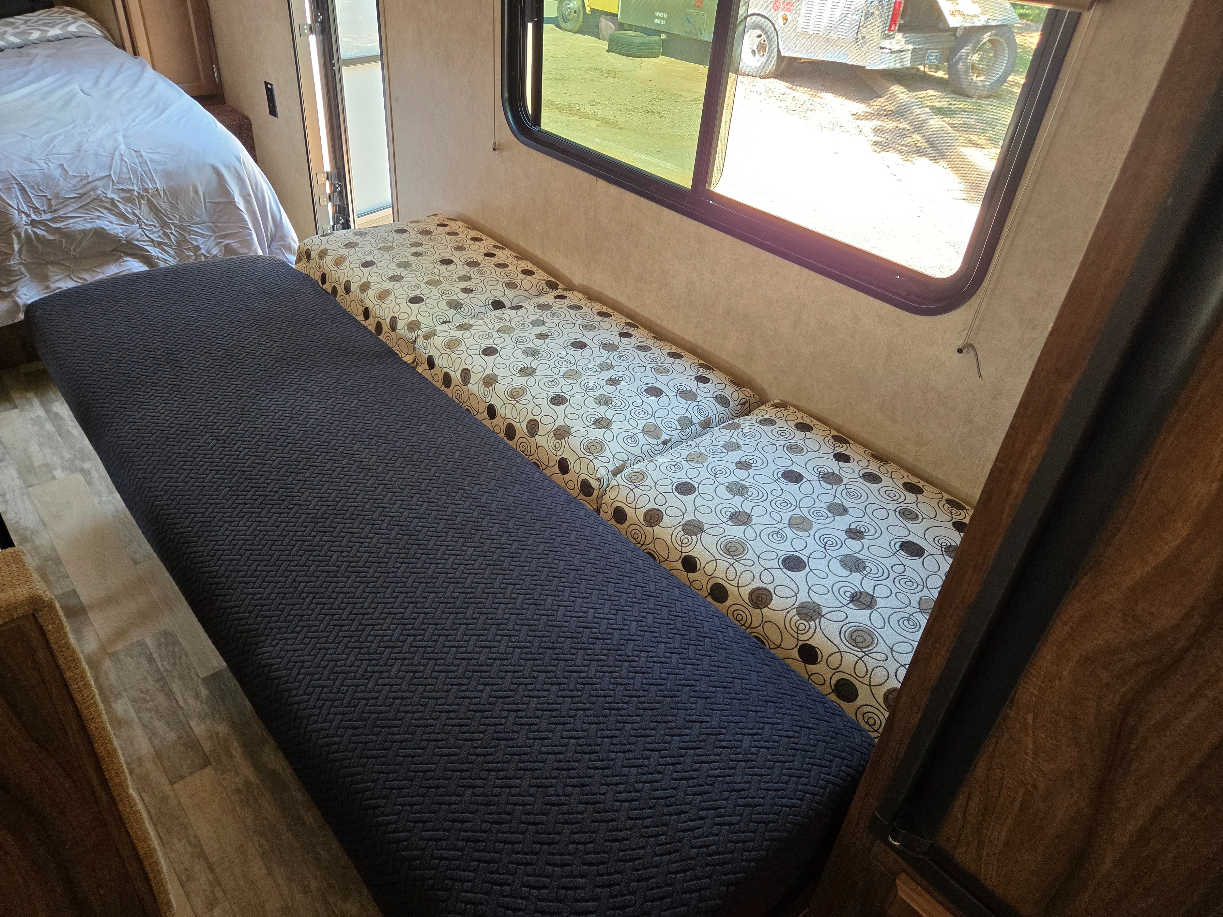 RV rental interior featuring a cozy bed, dark blue textured bench, and patterned dinette seating next to a window with an outdoor view.. Forest River Coachmen Classic 2018