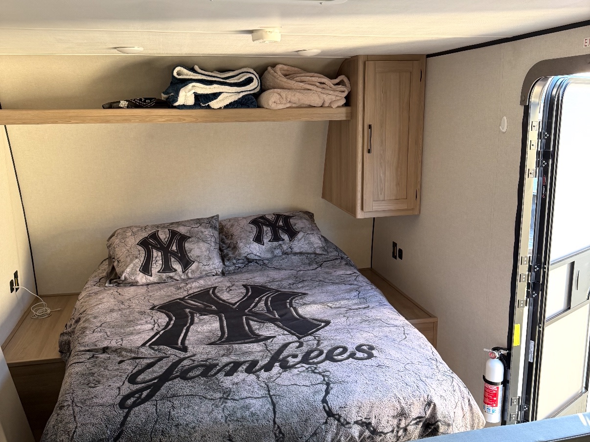 Cozy RV rental sleeping area with New York Yankees bedding, storage shelf, and cabinet. Fire extinguisher by the door.. Keystone RV Coleman 2025