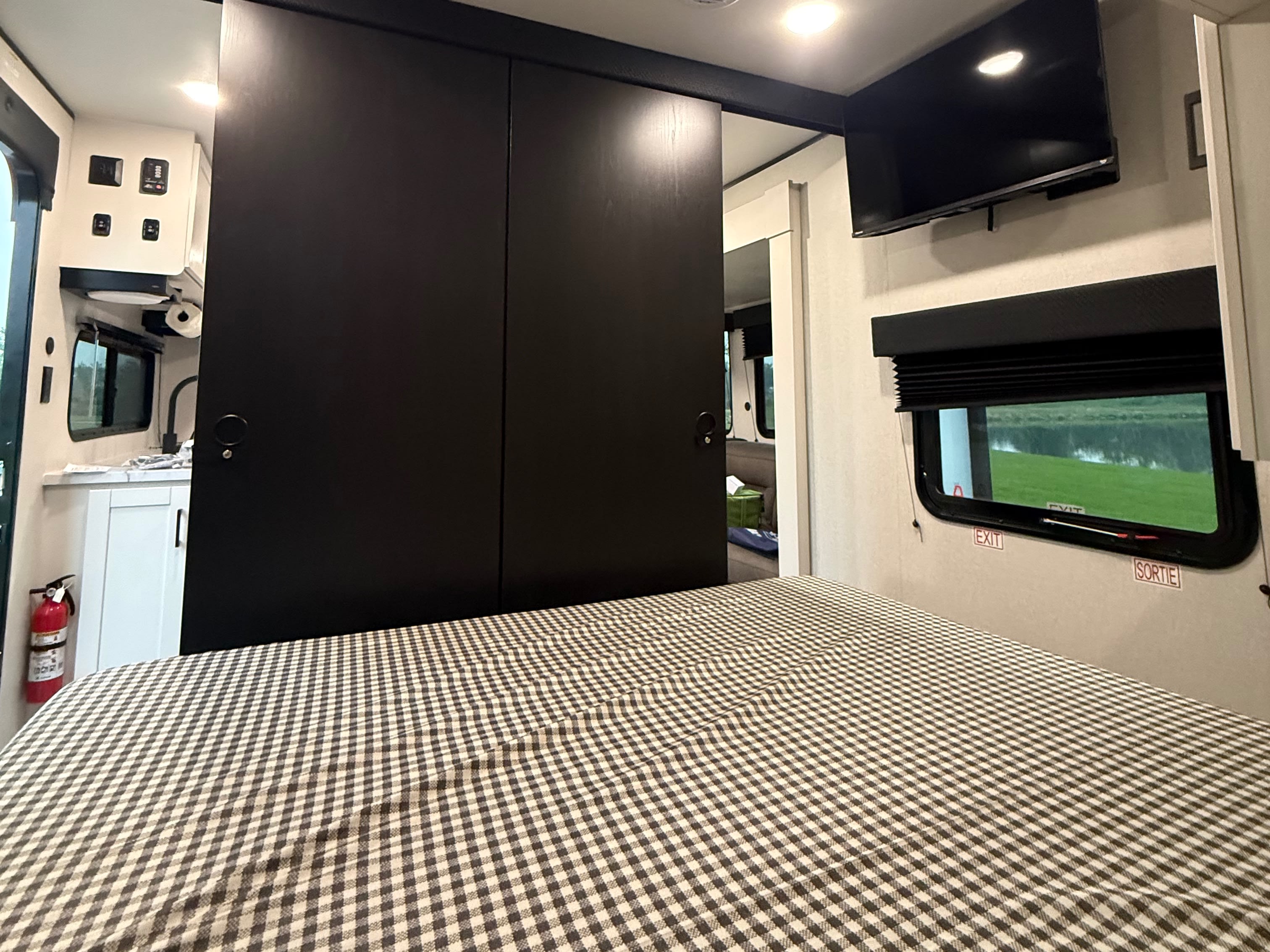RV rental interior: checkered bed, black sliding doors to a kitchen, TV, and window with an EXIT sign overlooking a lake.. Jayco Jay Flight SLX 2026