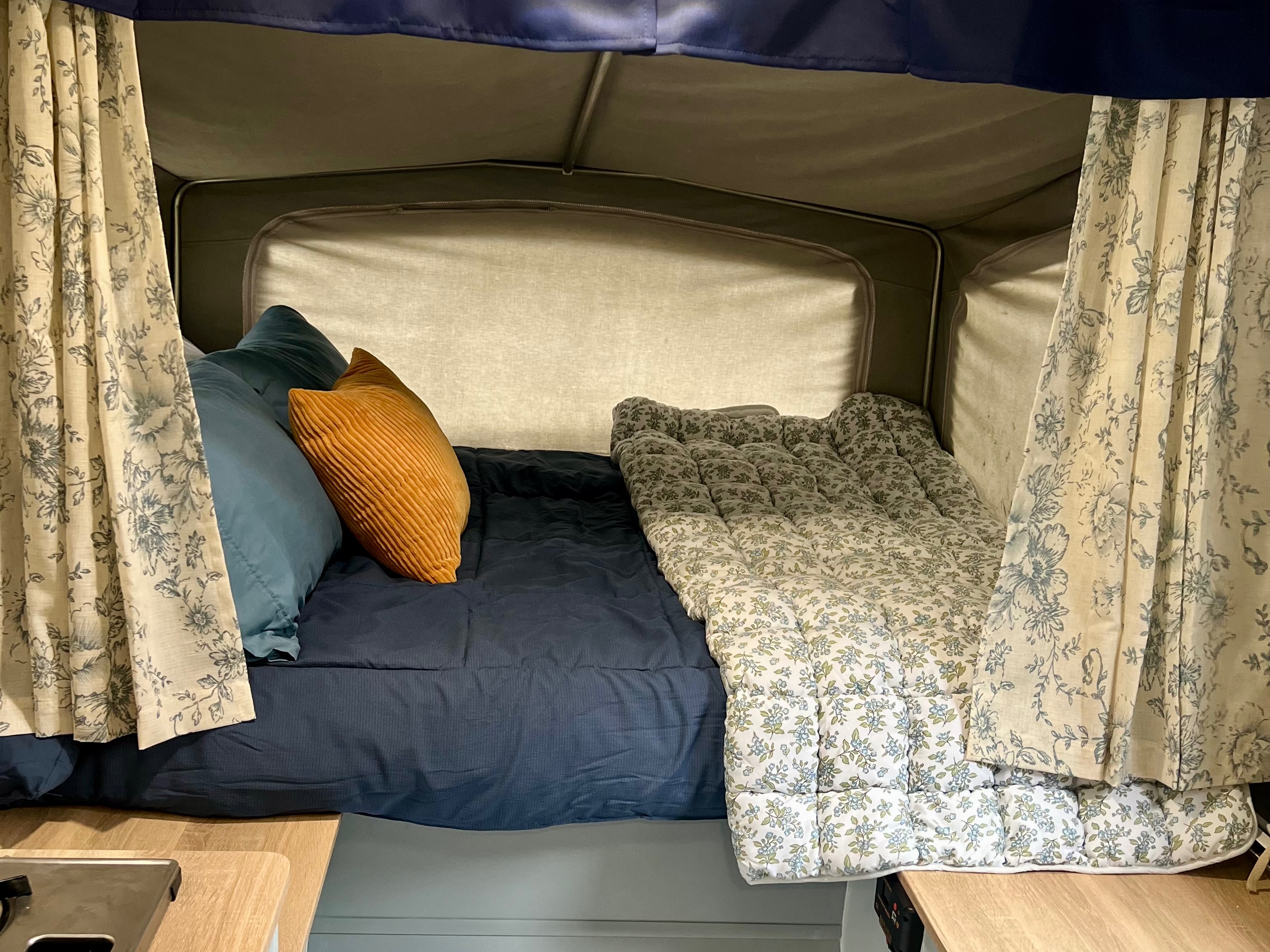 Cozy RV rental pop-up camper bed, featuring blue bedding, floral quilt, and patterned curtains. An inviting sleeping space.. Bonair BA650TT 2003