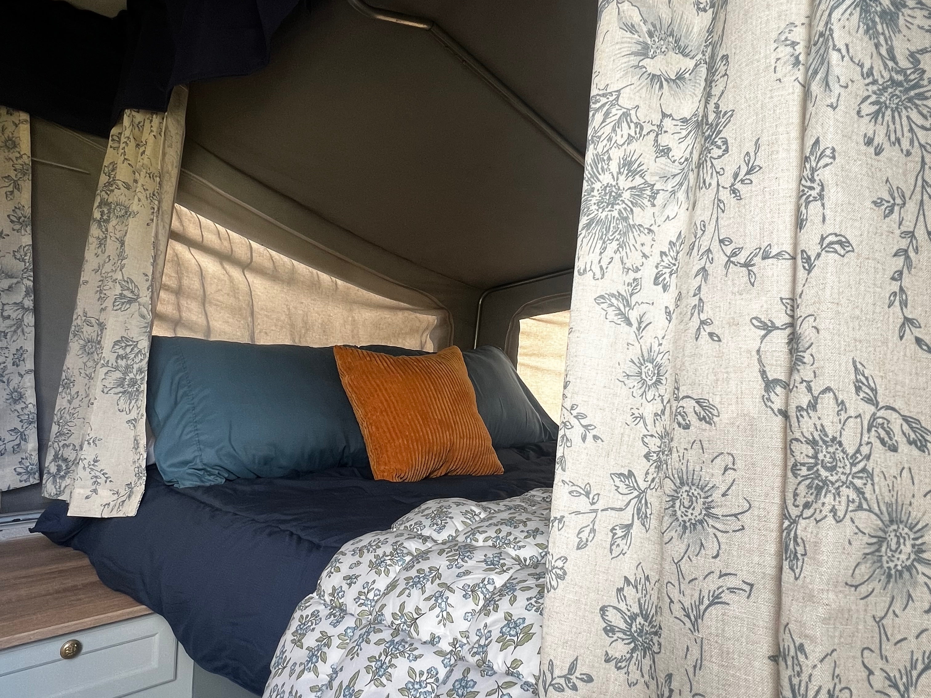 Experience a cozy RV rental: a comfortable bed with dark blue linens, an orange throw pillow, and charming floral curtains.. Bonair BA650TT 2003