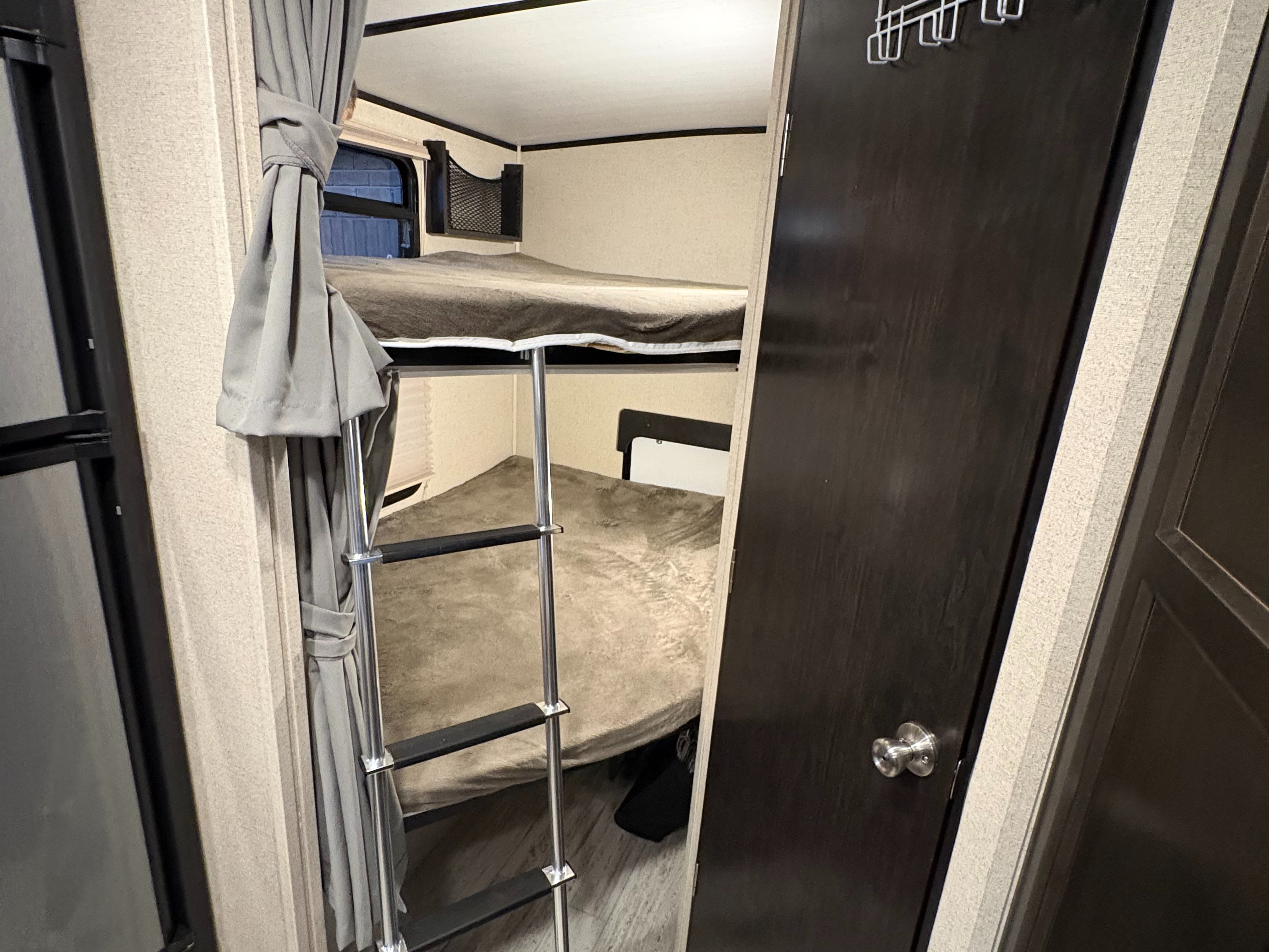 Cozy bunk beds with a ladder inside an RV rental, featuring grey curtains, a window, and a dark wood door.. Coachmen Northern Spirit 2454BH 2019