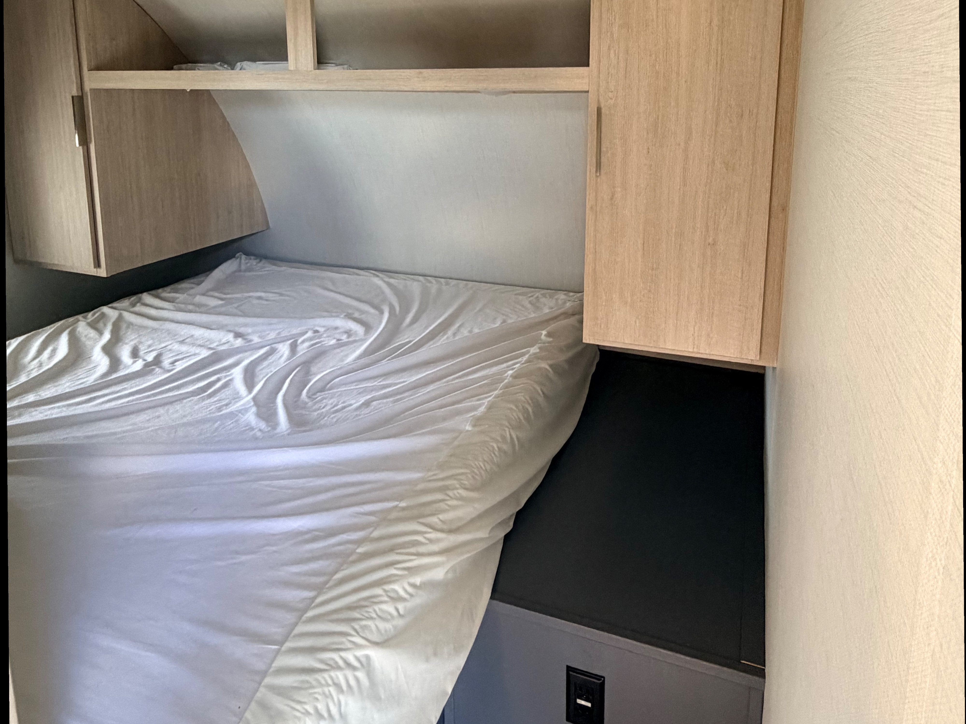 RV rental bedroom: a bed with white sheets, flanked by wooden overhead cabinets & shelves, offers compact sleeping space.. Forest River Tracer 2024