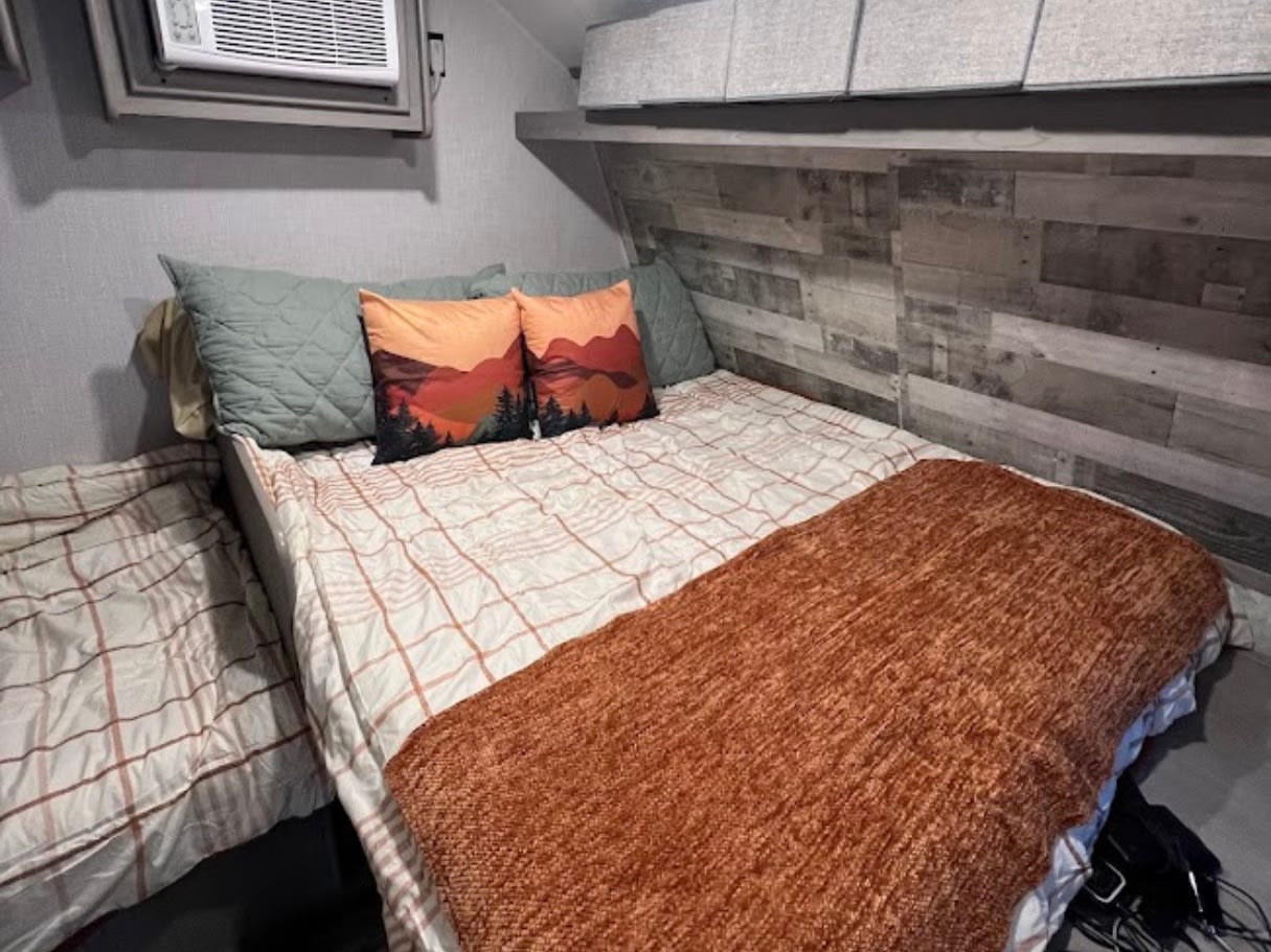 Comfortable RV rental sleeping area with AC, rustic wood accent wall, and cozy bedding. Mountain pillows add charm.. Dutchmen Coleman Lantern LT 2023
