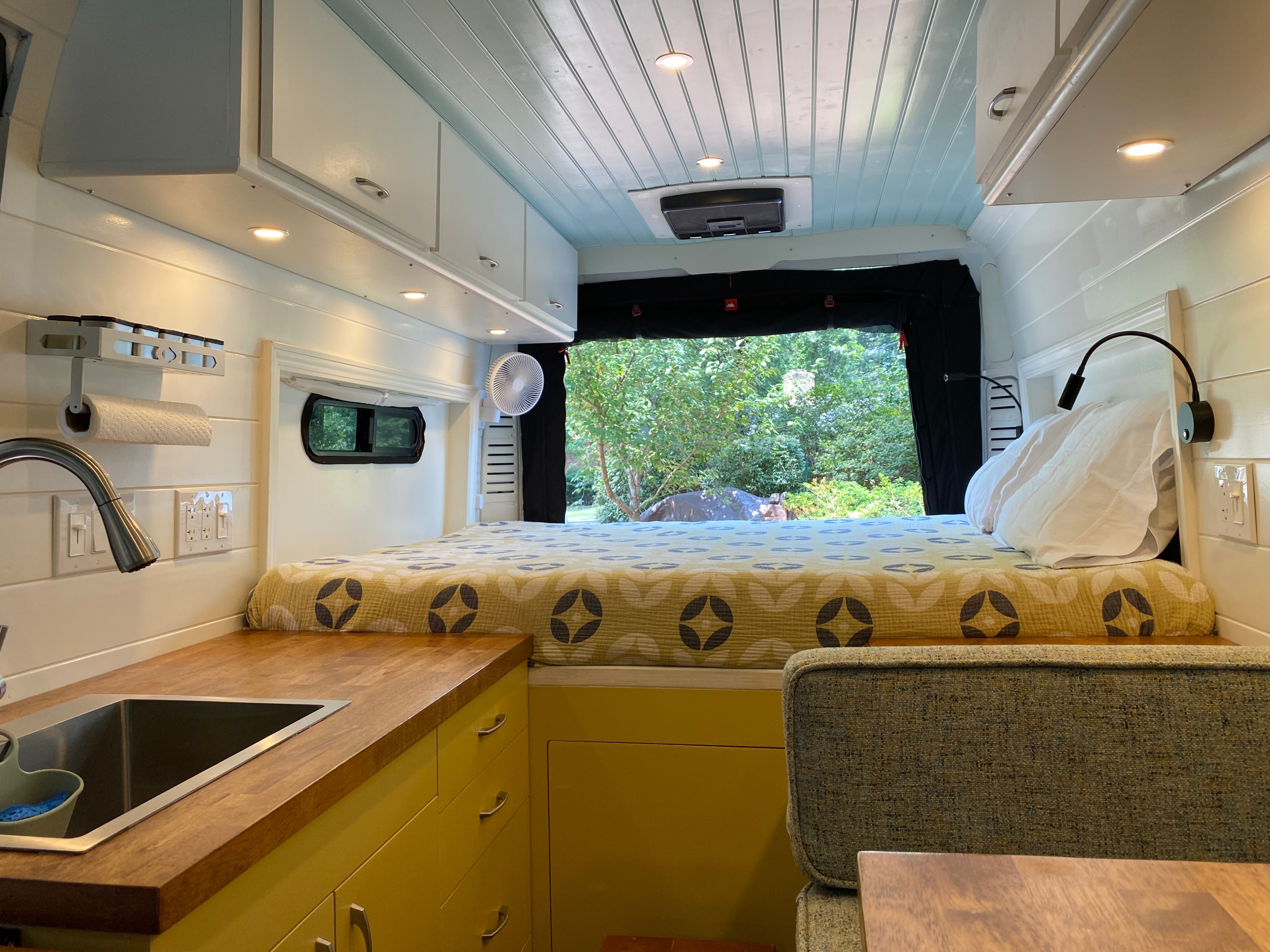 Cozy **rv rental** interior: a bed, built-in kitchenette with wood counter, overhead storage, and nature view outside.. Dodge Ram Promaster 3500 Hightop 2021