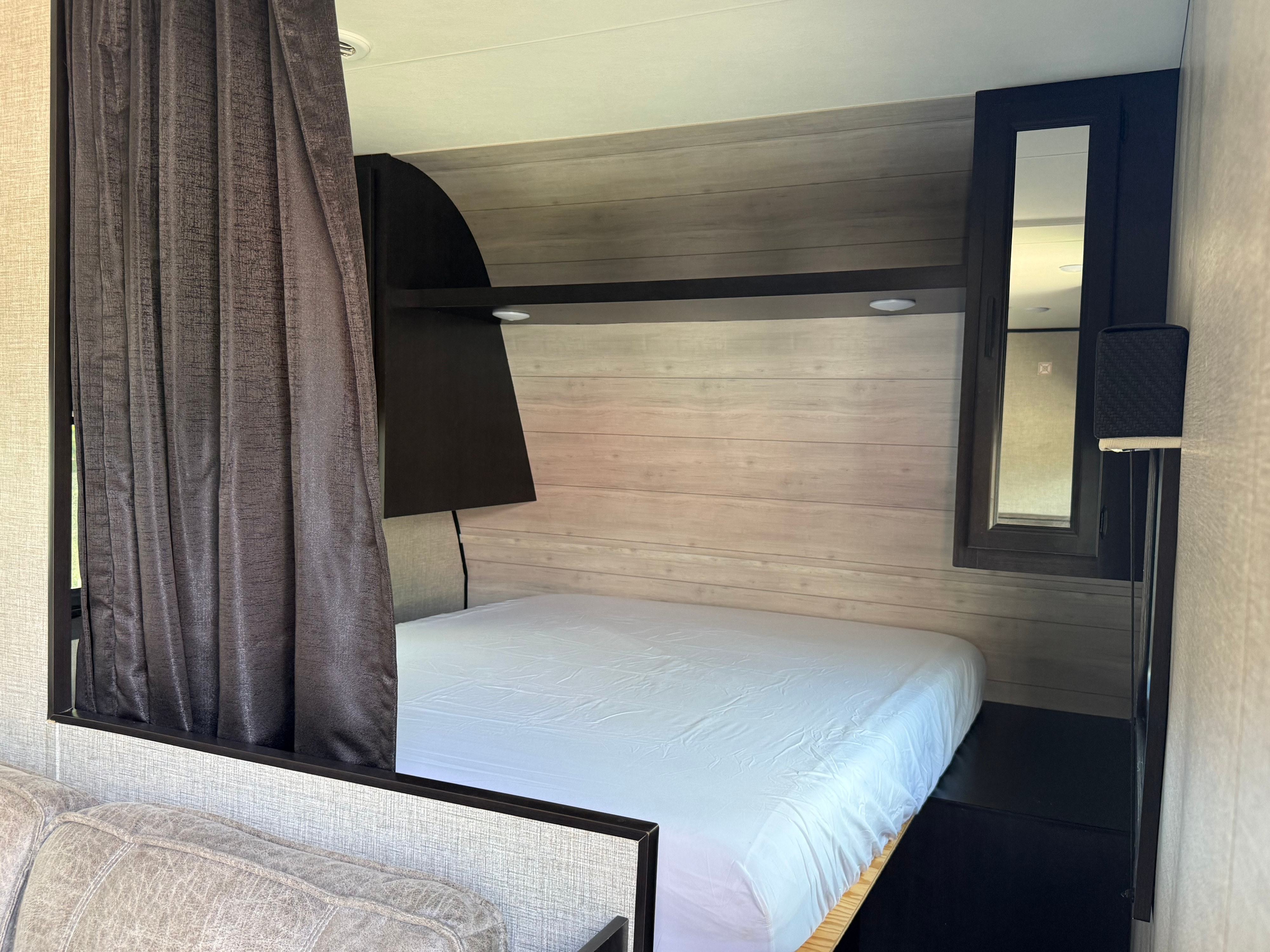 RV rental interior: cozy bed with white sheets, dark privacy curtain, wood-paneled wall, and lit overhead shelf.. Jayco Jay Flight SLX 2022