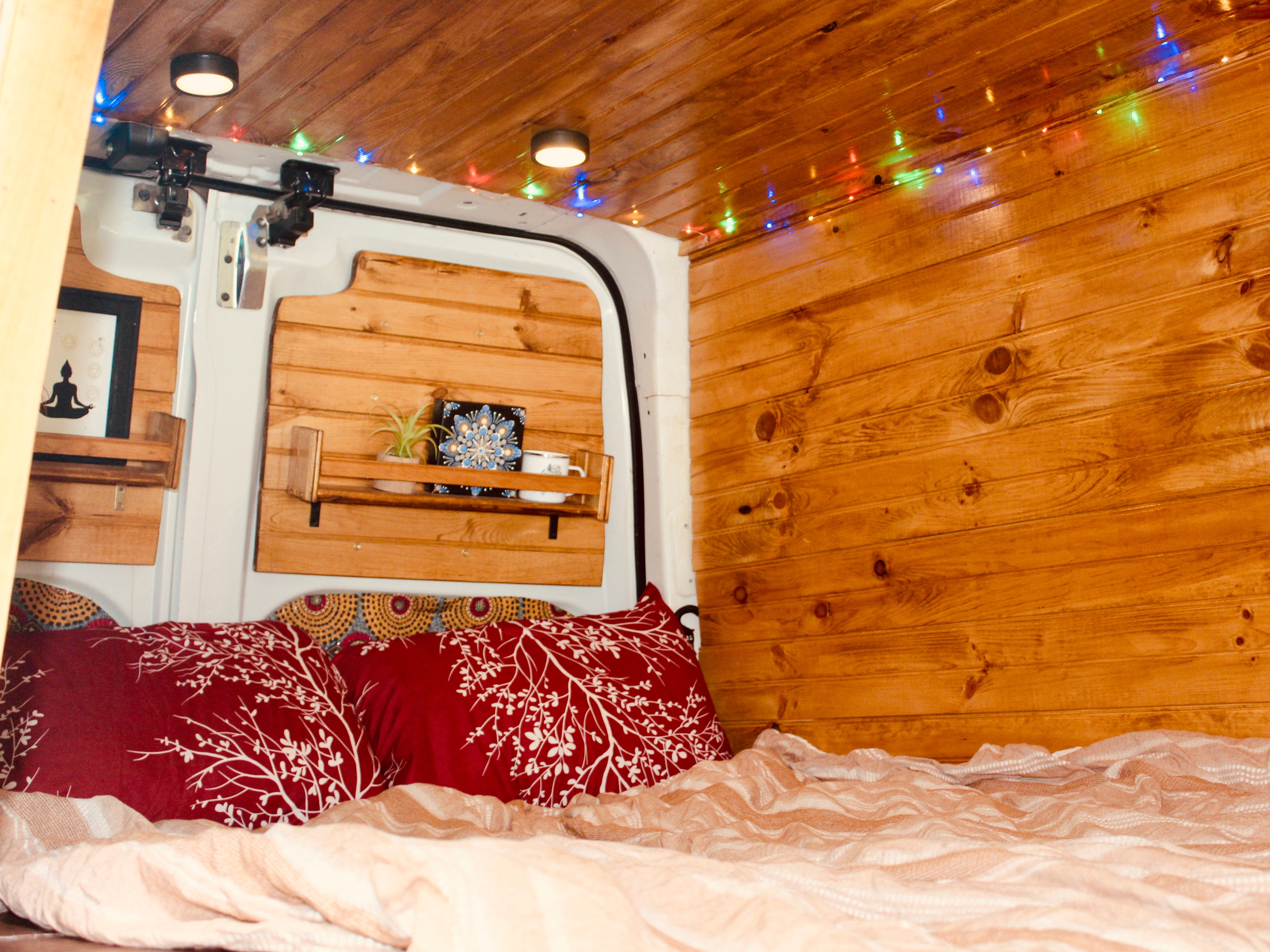 Cozy rv rental interior with wood paneling, festive string lights, and a comfy bed. Shelves hold decor like plants and art.. Nissan NV2500 2015