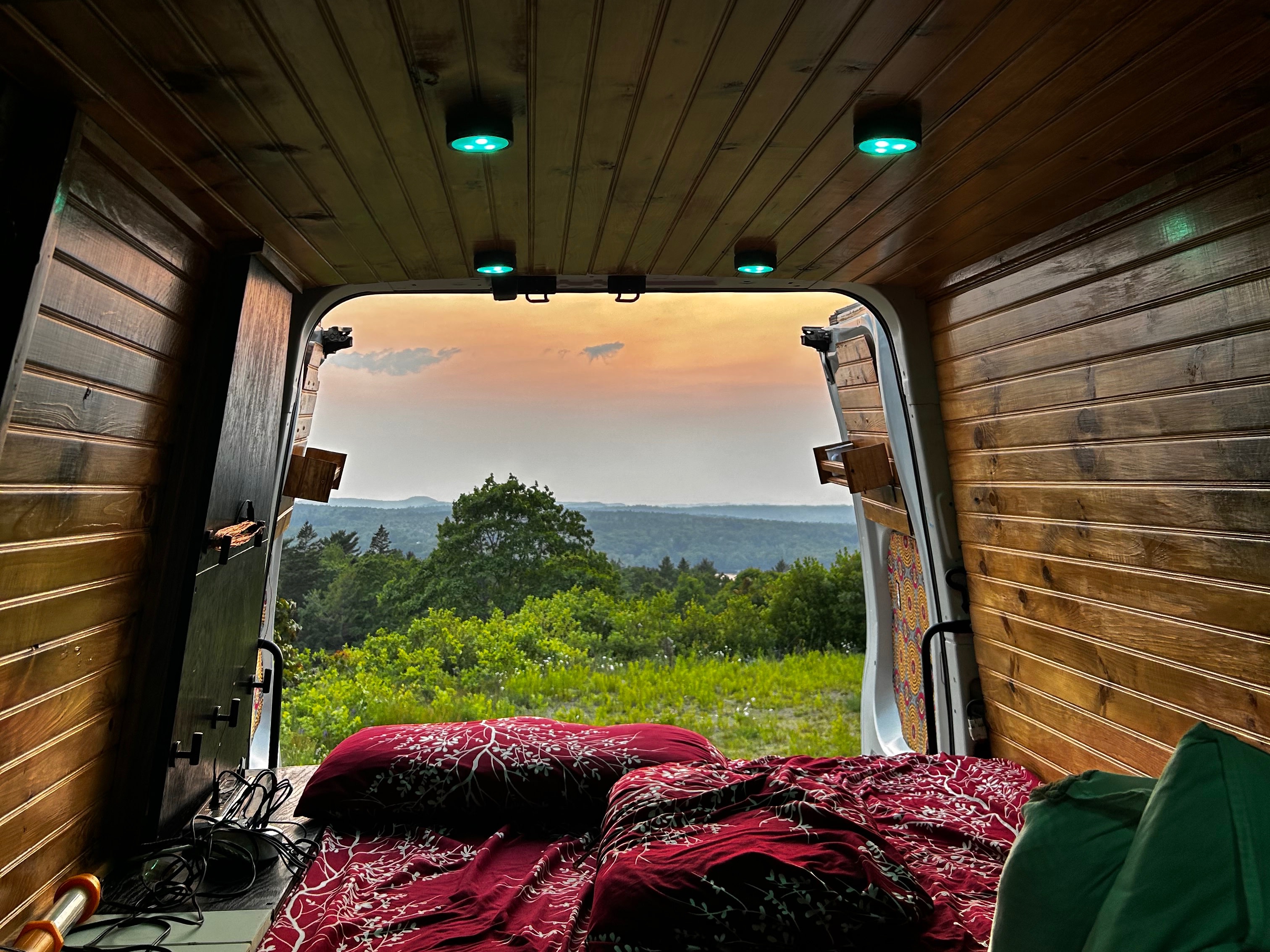 Inside a custom RV rental: wooden walls, red bedding, and green lights. Enjoy the panoramic sunset view over green hills.. Nissan NV2500 2015