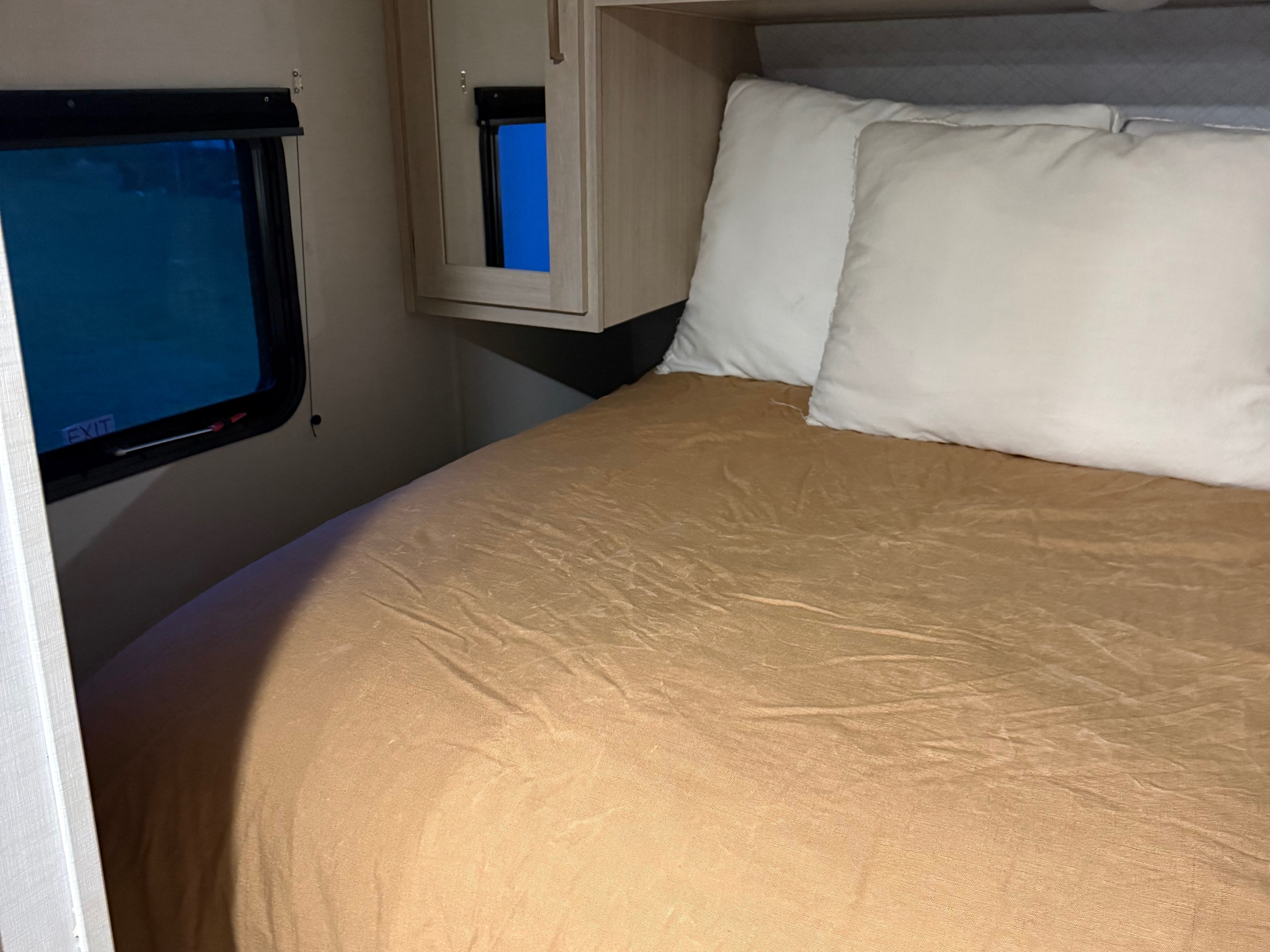 Comfortable RV rental bedroom featuring a bed, white pillows, cabinet, and window with an "EXIT" sign. Ideal for trips.. Avenger Avenger Trailer 2025