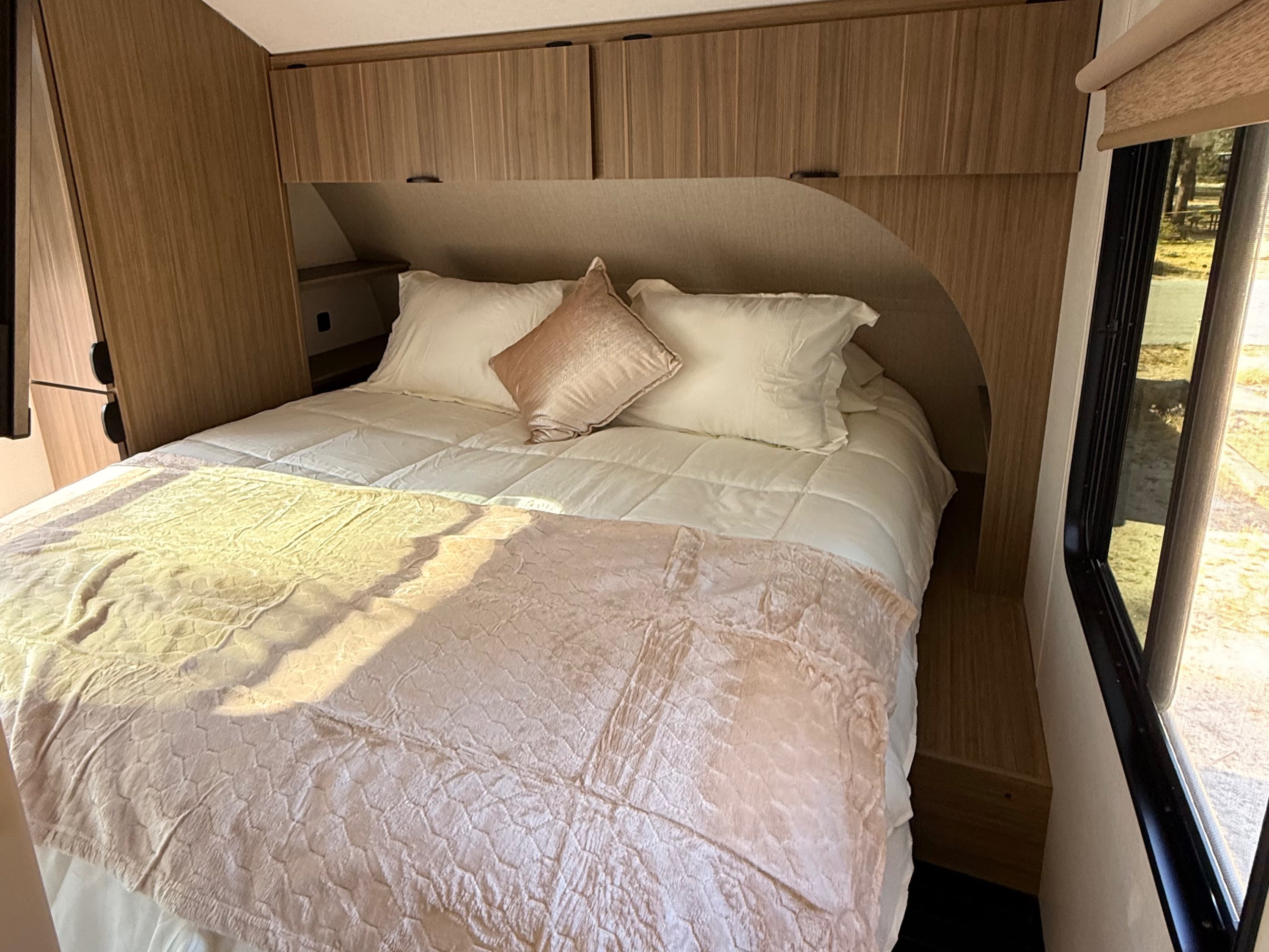 Cozy RV rental bedroom featuring a neatly made bed with white and pink linens, built-in wooden cabinets, and a sunlit window.. Forest River Wildwood X-Lite 2025