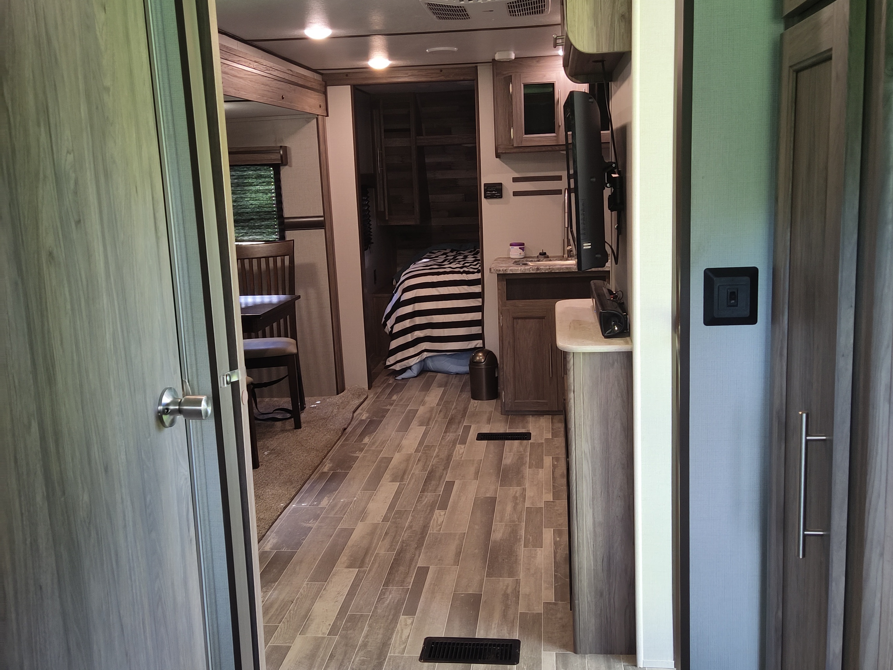 Interior of an inviting rv rental, showing a bedroom with striped bedding, a small kitchen, and a dining table. Modern RV design.. Zinger 280RB 2020