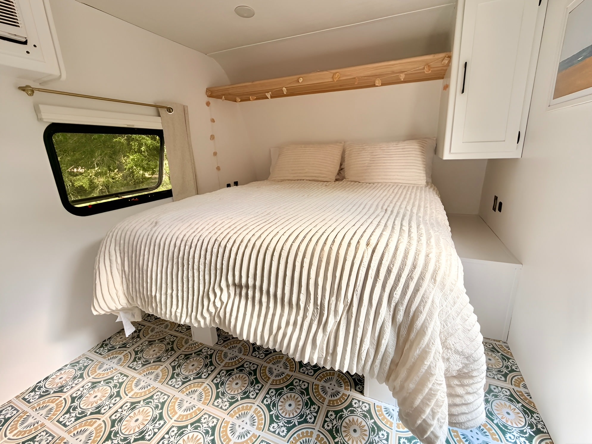 Bright RV rental bedroom featuring a plush bed, stylish patterned floor, window with greenery view, and overhead storage.. Keystone RV Coleman 2025