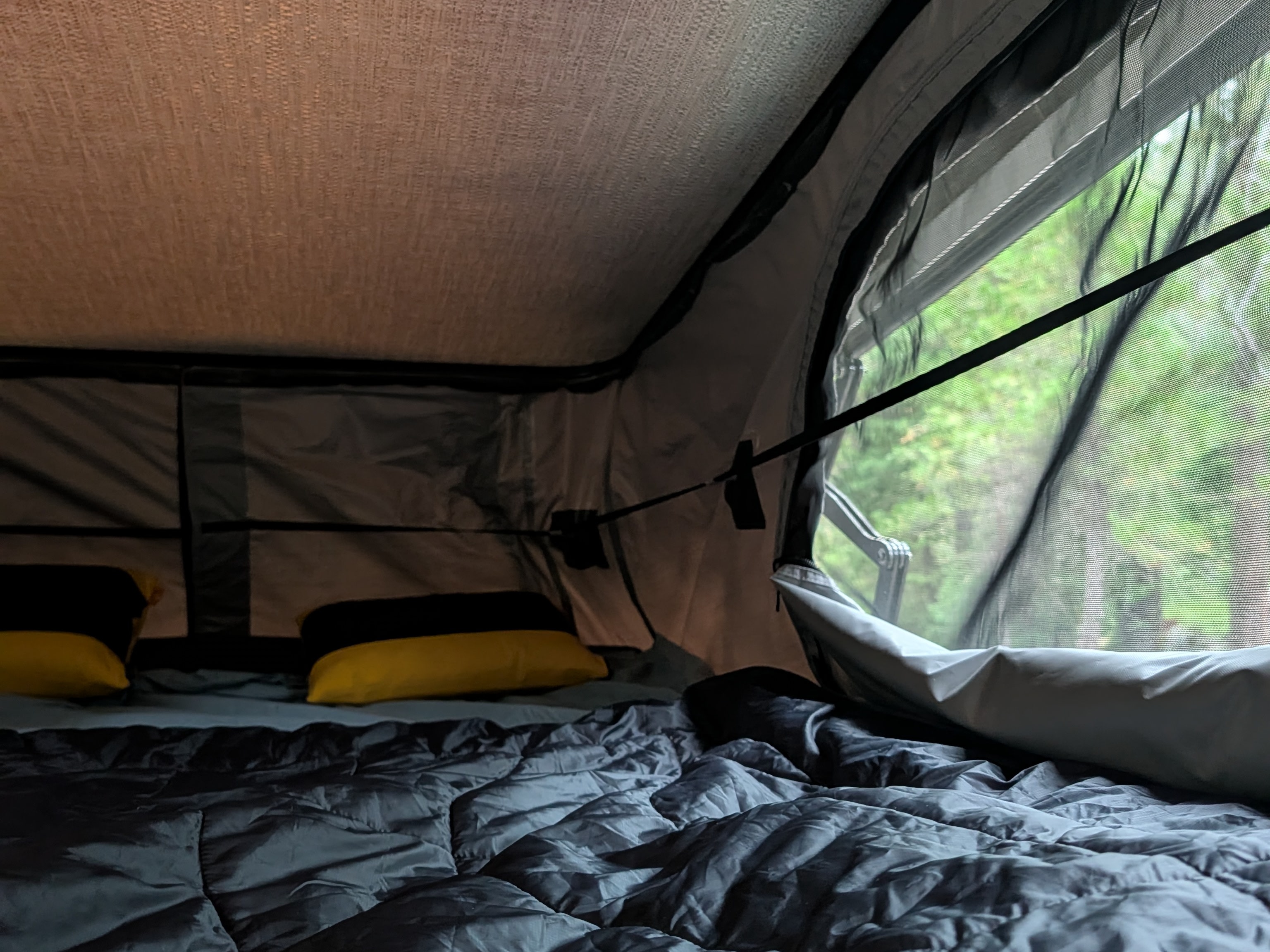 Cozy interior of an RV rental rooftop tent, featuring a bed with yellow pillows and a mesh window view of green trees.. Winnebago Solis 2025