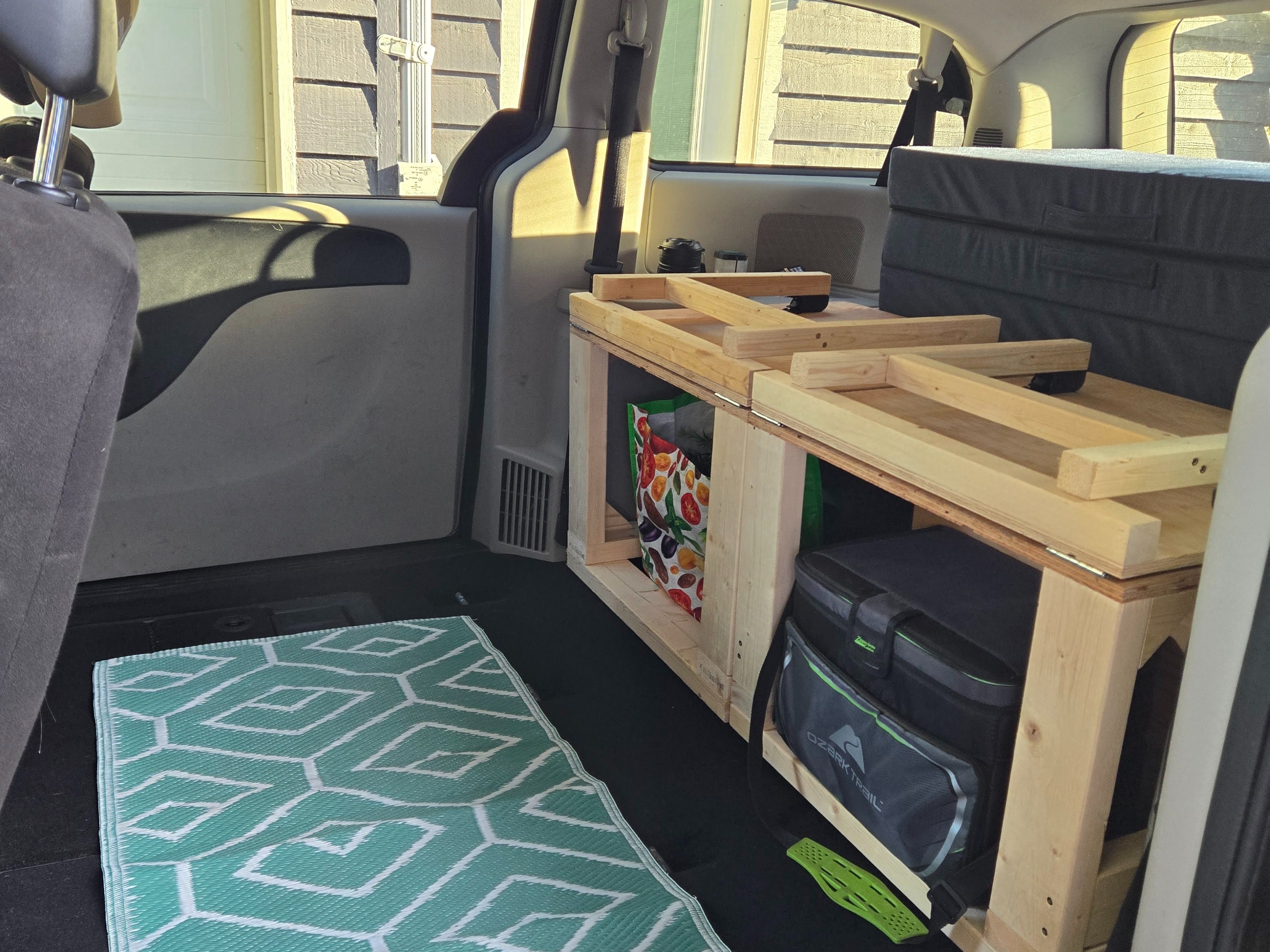 Custom car camping setup in a minivan, ideal for an rv rental. Features a wooden bed platform, cooler, and mat.. Dodge Grand Caravan 2014